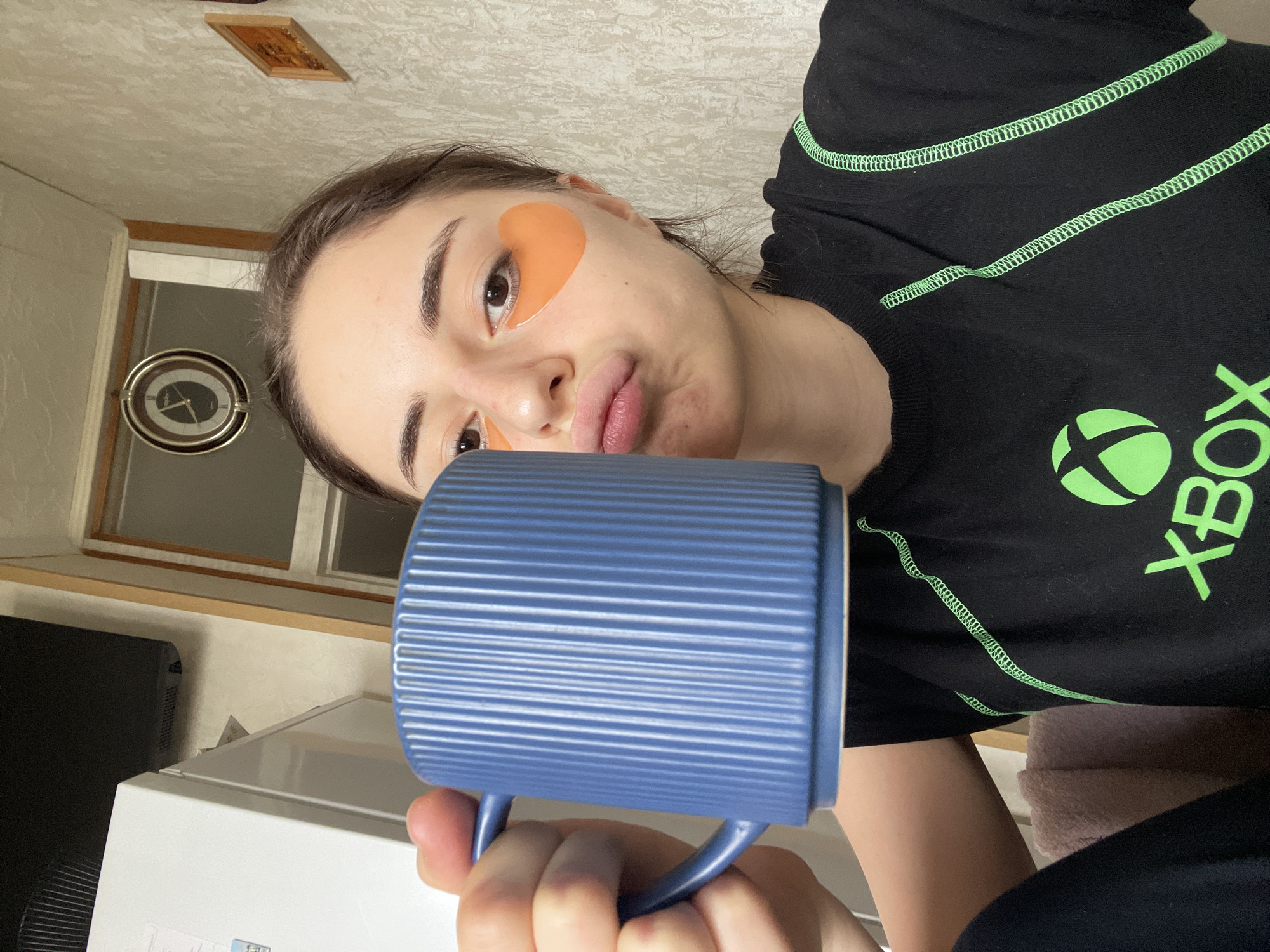 A young woman is holding a blue mug. She has orange eye patches under her eyes. She is wearing a black t-shirt with a green Xbox logo. There is a clock on the wall behind her. The location is not discernable as Moscow, Russia, and there are no identifiers of any city or region.