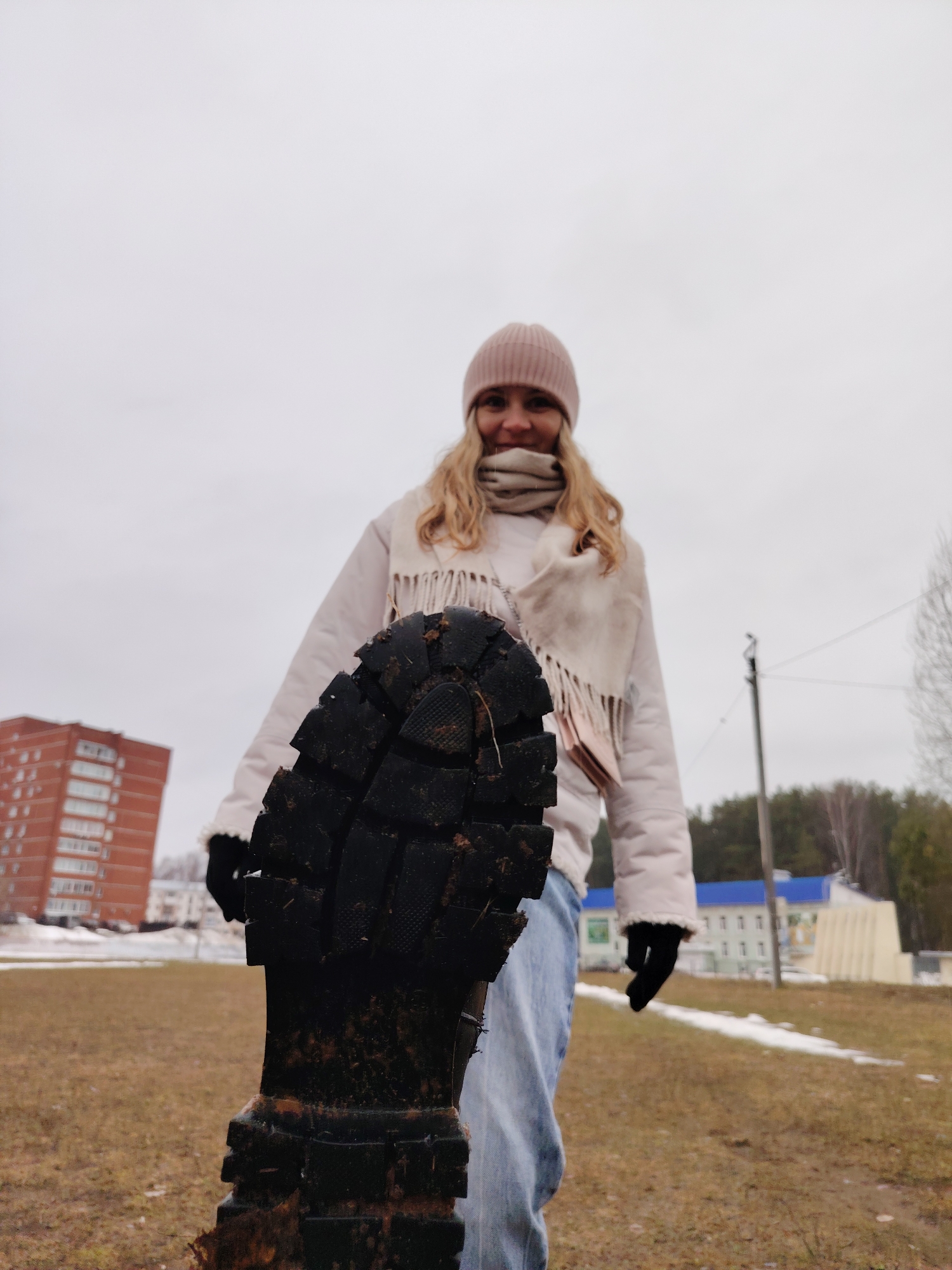 The image shows a woman standing in a grassy area in what appears to be the city of Chaikovsky, Russia. She is wearing a pink hat, a light-colored scarf, a white jacket, blue jeans, and black gloves. The focal point is a muddy, black boot sole positioned close to the camera. In the background, there is a red brick apartment building on the left, and a building with a blue roof further back on the right, along with some trees and a utility pole. Patches of snow remain on the ground, suggesting early spring or late autumn. The sky is overcast. The woman is looking towards the camera with a slight smile.