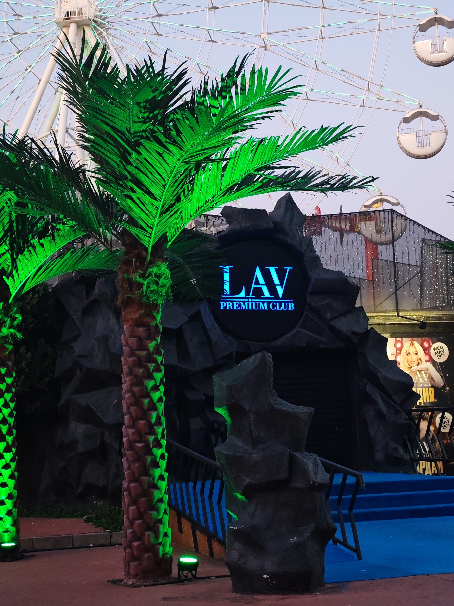 The image depicts the entrance of "LAV Premium Club" in Nesebar, Bulgaria. The club's entrance is stylized as a dark, jagged rock formation with a blue neon sign displaying the club's name. Two artificial palm trees, illuminated with green light, flank the entrance. A Ferris wheel is visible in the background, suggesting the club is located in an entertainment district. Blue steps lead up to the entrance, and a smaller rock formation acts as a decorative element nearby. Posters and lights are also visible, indicating an active nightlife environment.