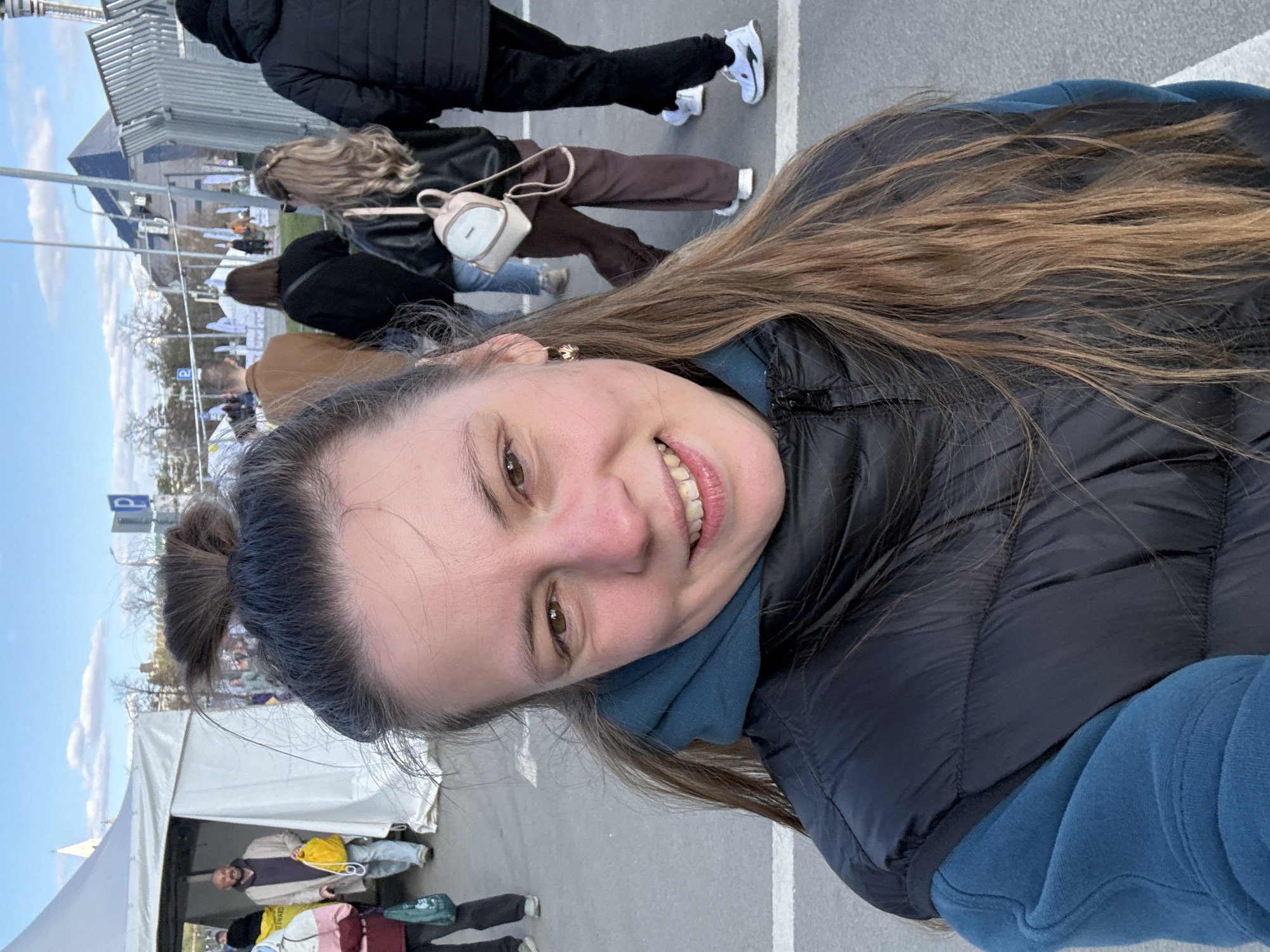 A selfie photograph depicts a woman outdoors in Kazan, Russia. She is smiling and wearing a blue hooded sweatshirt and a black puffer vest. Her hair is partially pulled up in a bun. In the background, multiple people are walking on a paved area. There is a white tent visible to the left, and a building complex in the distance. Parking space signage is present in the midground. The weather appears to be clear and sunny.
