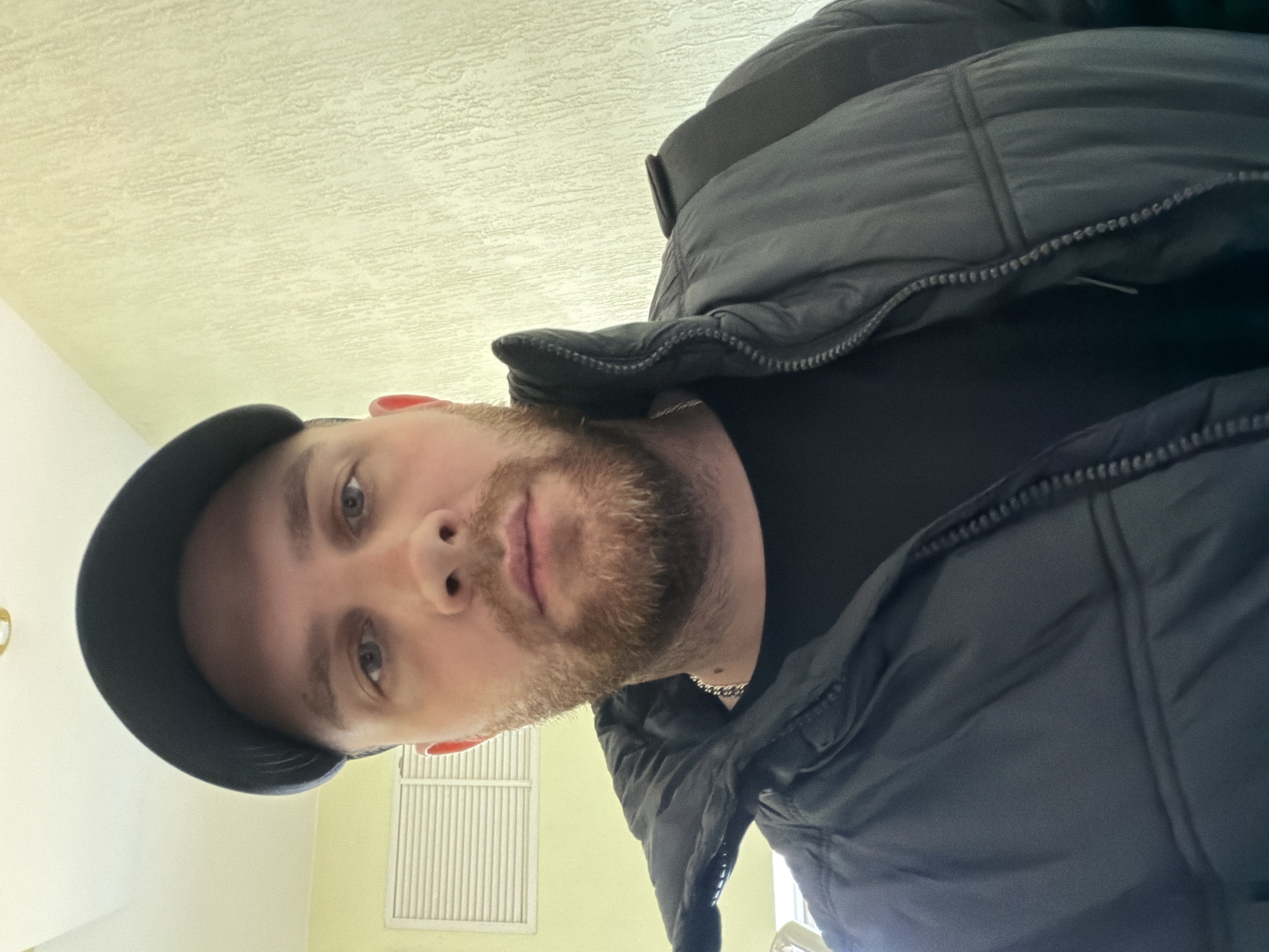 A man with a beard and a black cap is wearing a black puffer jacket and looking down at the camera. He appears to be indoors, with a light yellow wall behind him. A white vent is visible in the lower left corner of the image. The location is not evident from the image.