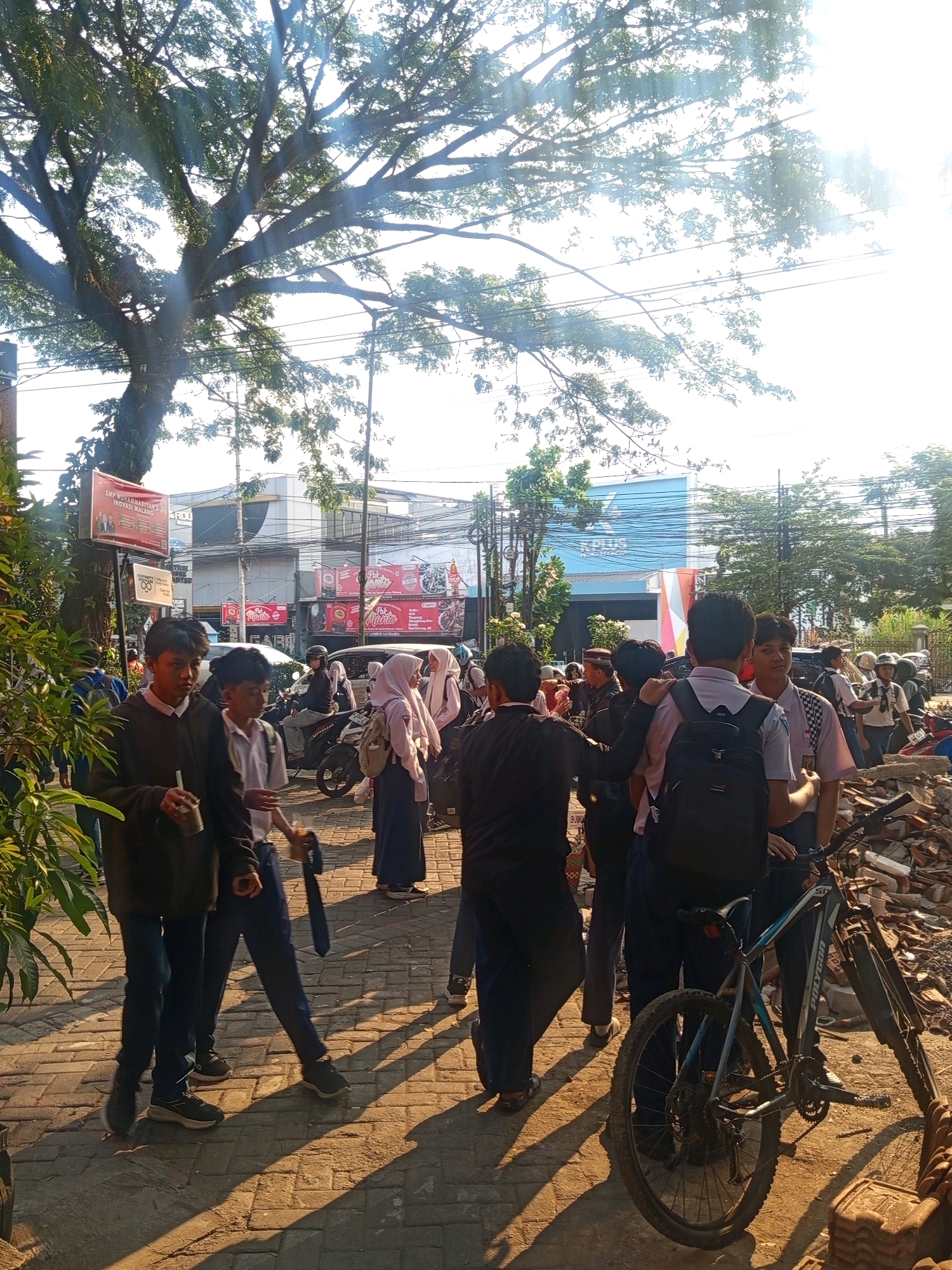 This image captures a street scene in Malang, Indonesia, likely near a school. A group of young people, predominantly dressed in school uniforms consisting of white shirts and dark trousers or skirts, are visible. Many appear to be students, with some carrying backpacks. Sunlight streams through a large tree, casting long shadows across the paved walkway. In the background, buildings with storefronts are present, including one with a large blue sign. Several motorcycles are parked or being ridden, indicating typical urban traffic. A bicycle is leaning against a pile of debris on the right side of the image. The overall atmosphere suggests a bustling, everyday moment in a populated urban area.