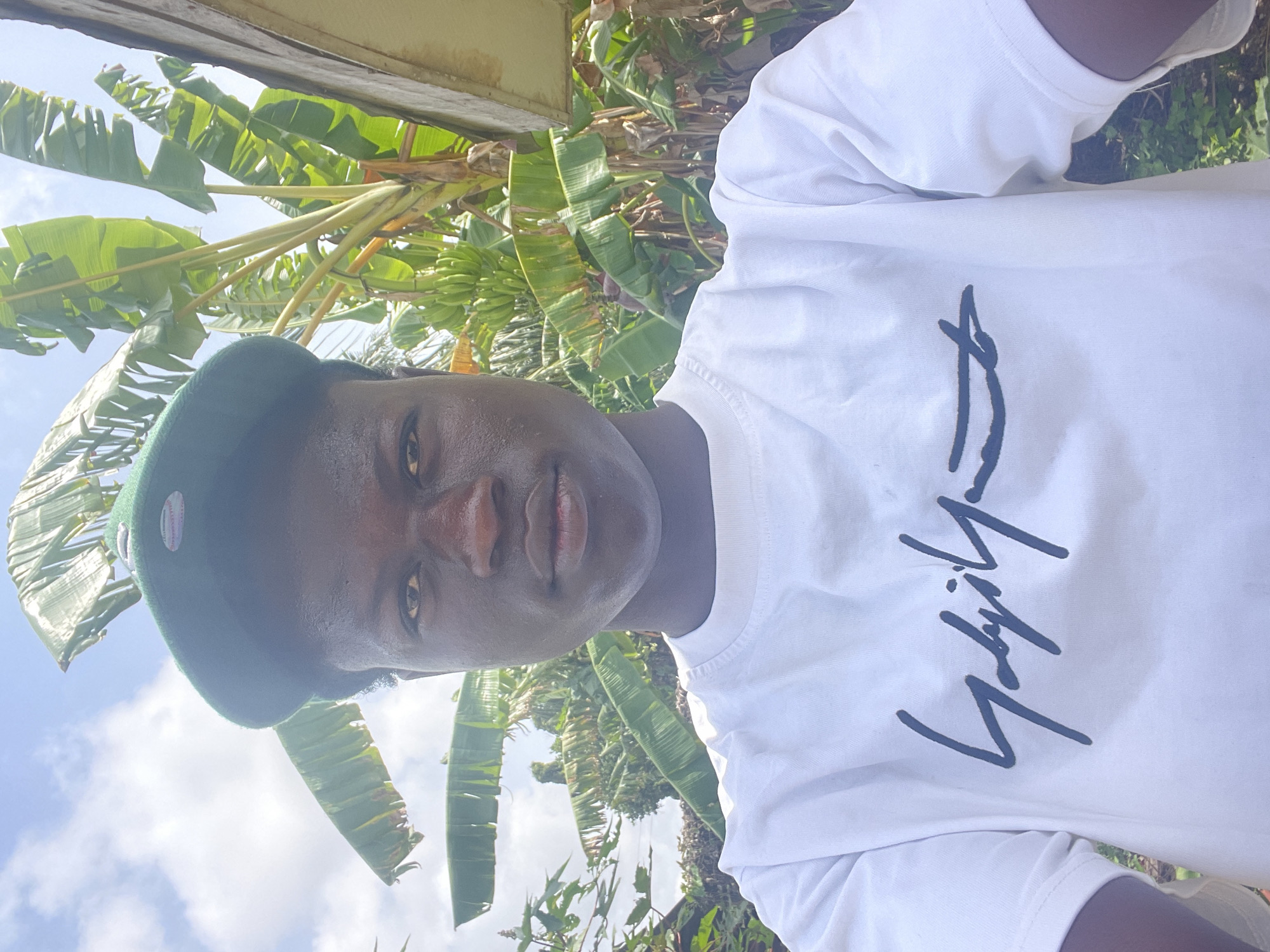 A young man is depicted in a selfie taken outdoors in Badagry, Nigeria. He wears a white t-shirt with a signature-style graphic and a green beret. The background features banana trees and glimpses of a cloudy sky.