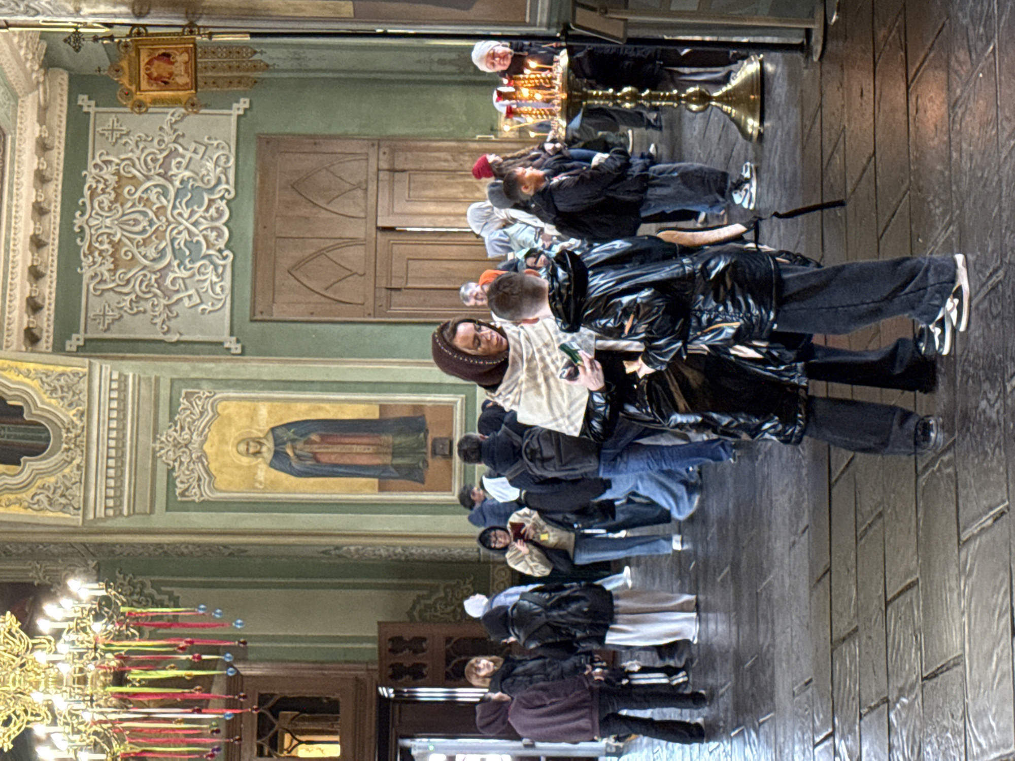 The image shows the interior of a building, possibly a church or cathedral, in Kazan, Russia. Several people are present, some standing and others moving within the space. The environment is ornate, with detailed wall paintings and architectural elements. There's a large chandelier with colorful decorations hanging from the ceiling. A woman in a brown headscarf appears to be taking a photograph with a mobile phone while standing next to a person in a shiny black jacket. There are several rows of people congregating near a candle stand. The floor is made of dark, possibly wooden, planks.