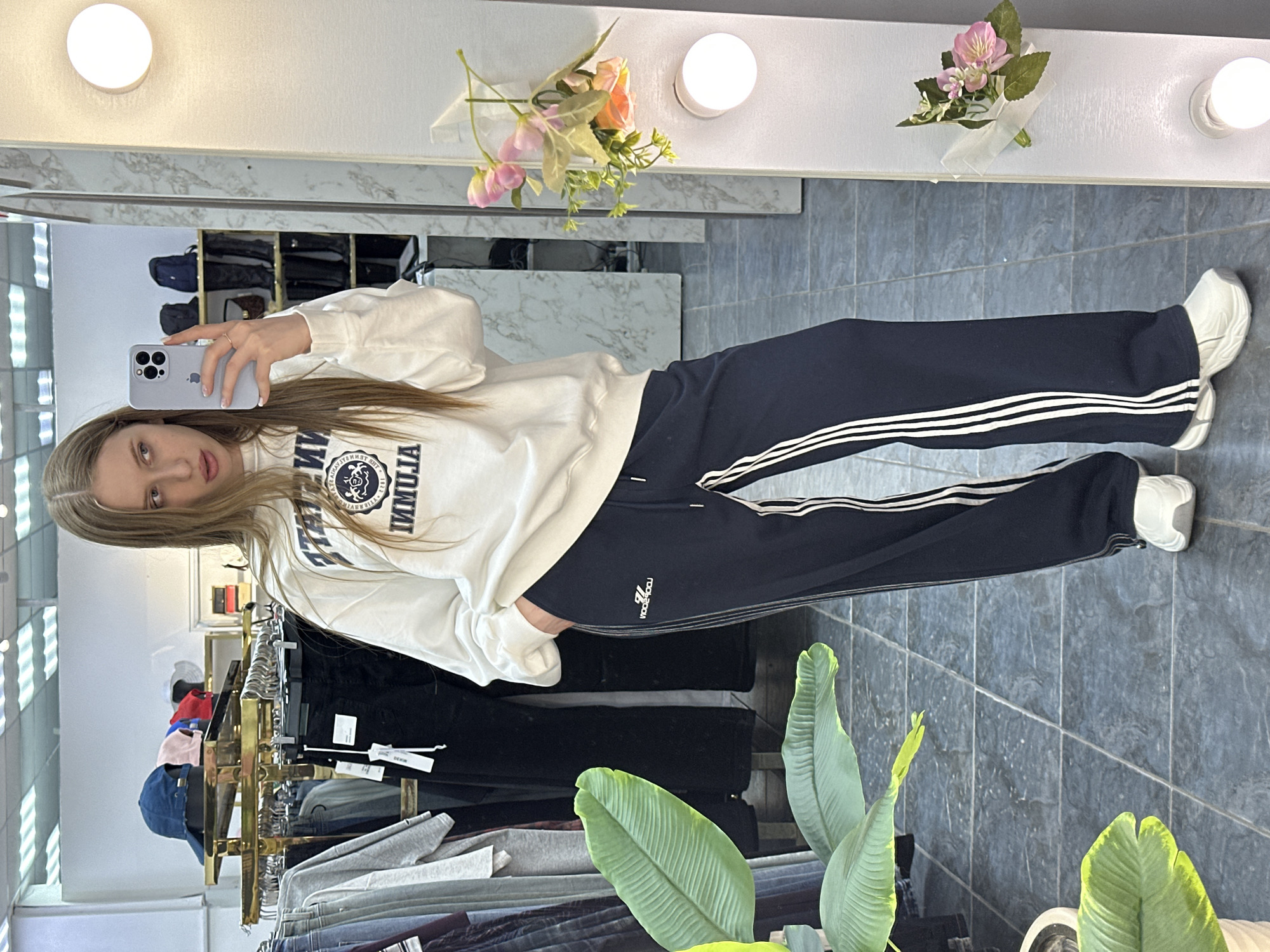 A woman with long blonde hair is taking a selfie in front of a mirror. She is wearing a white sweatshirt with a dark blue logo and dark blue sweatpants with white stripes on the sides. She is wearing white sneakers. Behind her, there are clothing racks with various items of clothing. In the foreground, there is a potted plant with large green leaves. The mirror is in a retail setting, likely a clothing store. The location appears to be in the city of Chaikovsky, Russia.