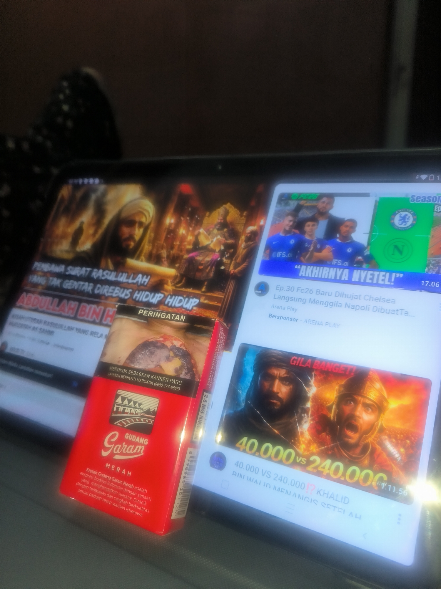 The image shows a red pack of "Gudang Garam Merah" cigarettes placed on a tablet displaying videos. The tablet's screen shows content including a video about "PEMBAWA SURAT RASULULLAH YANG TAK GENTAR DIREBUS HIDUP HIDUP ABDULLAH BIN H" (a messenger of the Prophet Muhammad who was not afraid to be boiled alive, Abdullah bin H), a video about Chelsea football and a video titled "40,000 vs 240,000" about a battle and mentions a person named Khalid.