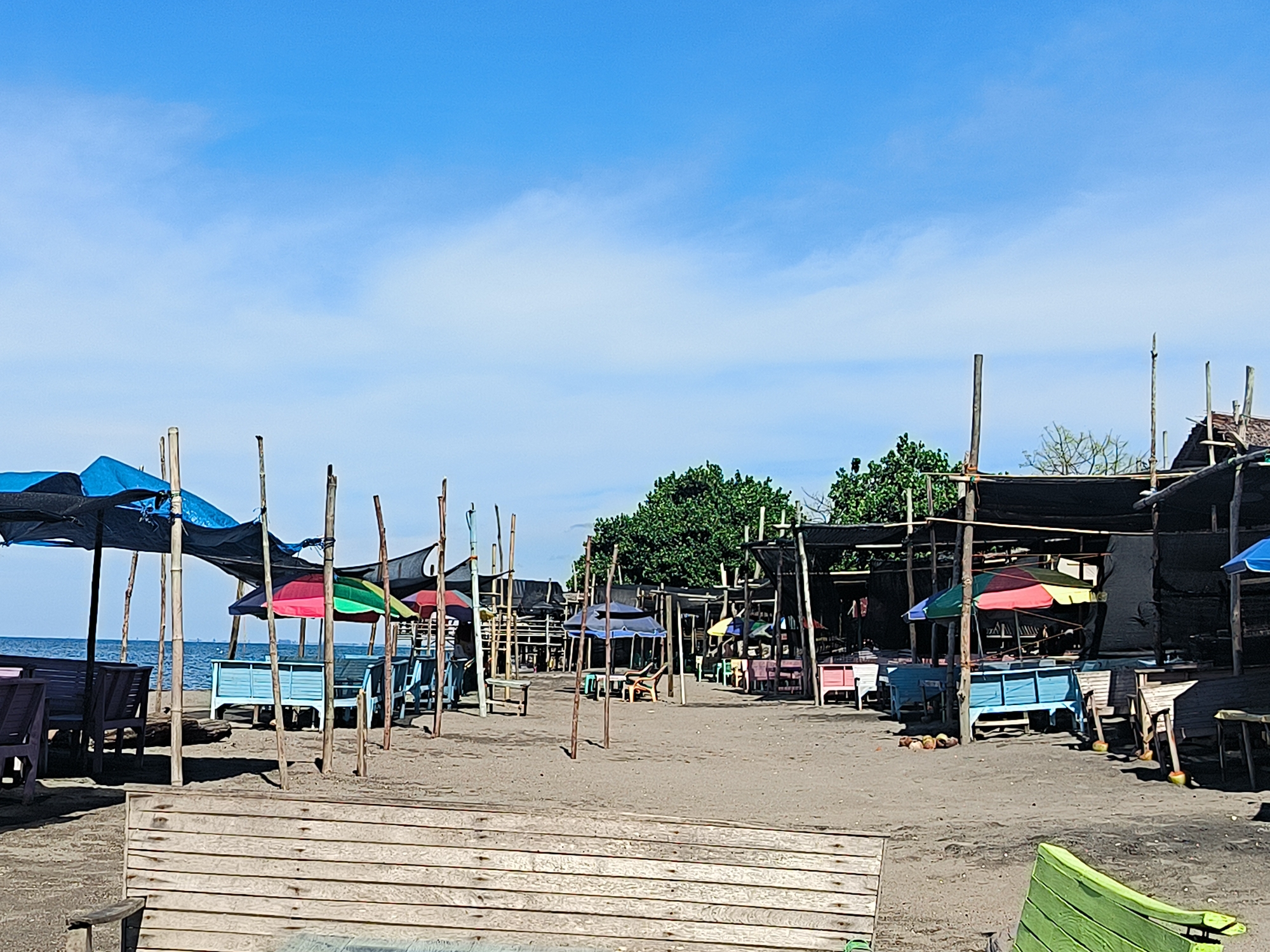 The image depicts a beach scene in Lhokseumawe, Indonesia. The location appears to be a local beach area with simple structures providing shade, likely small restaurants or stalls.
**Environment:** The ground is sandy. The sky is blue with light clouds.
**Objects:** Several makeshift structures with wooden supports and fabric or netting roofs are visible. Colorful umbrellas provide additional shade. Benches and tables, painted in various colors, are arranged under the shelters. Coconuts lie scattered on the sand.
**People:** There are no individuals visible in the image.
**Subjects:** The focus is on the beach stalls.
**Actions:** The scene suggests a place for leisure and food service, though no activity is actively occurring at the time of the picture.
**Interactions:** Without visible people, there are no observable interactions taking place.