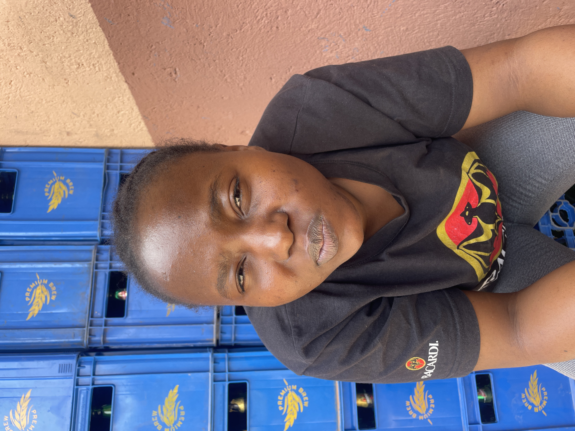 The image features an adult person, likely female, with dark skin and short, dark, natural hair, looking directly at the viewer with a neutral expression. They are wearing a black short-sleeved t-shirt with a colorful graphic design on the chest (red, yellow, and black elements) and a "BACARDI." logo visible on the right sleeve.
The person is positioned against a background of stacked blue plastic crates. Many of these crates have the text "PREMIUM BREW" in yellow, along with a wheat or barley symbol, indicating they are likely for beverages such as beer. Bottles can be seen inside some of the crates. The background also includes a wall painted in two shades of brown, with a distinct diagonal line separating a lighter and darker section. The bright lighting suggests it is daytime. The overall setting appears to be an outdoor or semi-open commercial area, possibly a beverage distribution point or shop, located in Jos, Nigeria.