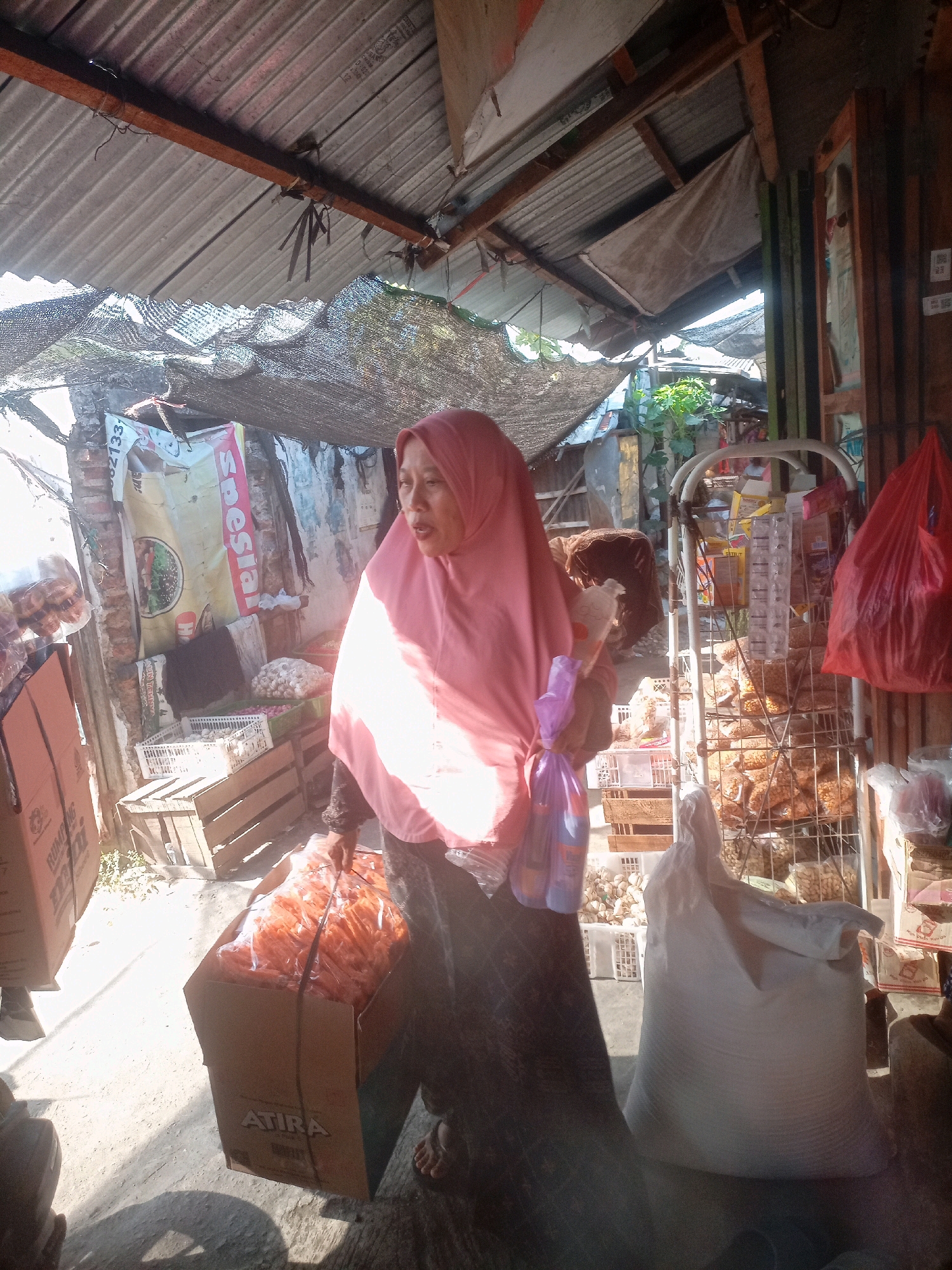 The image depicts an outdoor market scene in Sluke, Indonesia, under a corrugated metal roof and black netting overhead. A woman wearing a pink hijab and a dark patterned dress stands in the foreground, holding a clear plastic bag containing orange-colored items and a brown box with the "ATIRA" logo. She is looking to her right, with an open mouth, as if speaking. Beside her is a large white sack. In the background, a metal rack is filled with packaged snacks. Wooden crates and other market goods are visible, along with a red plastic bag hanging on the right. The ground appears to be paved.