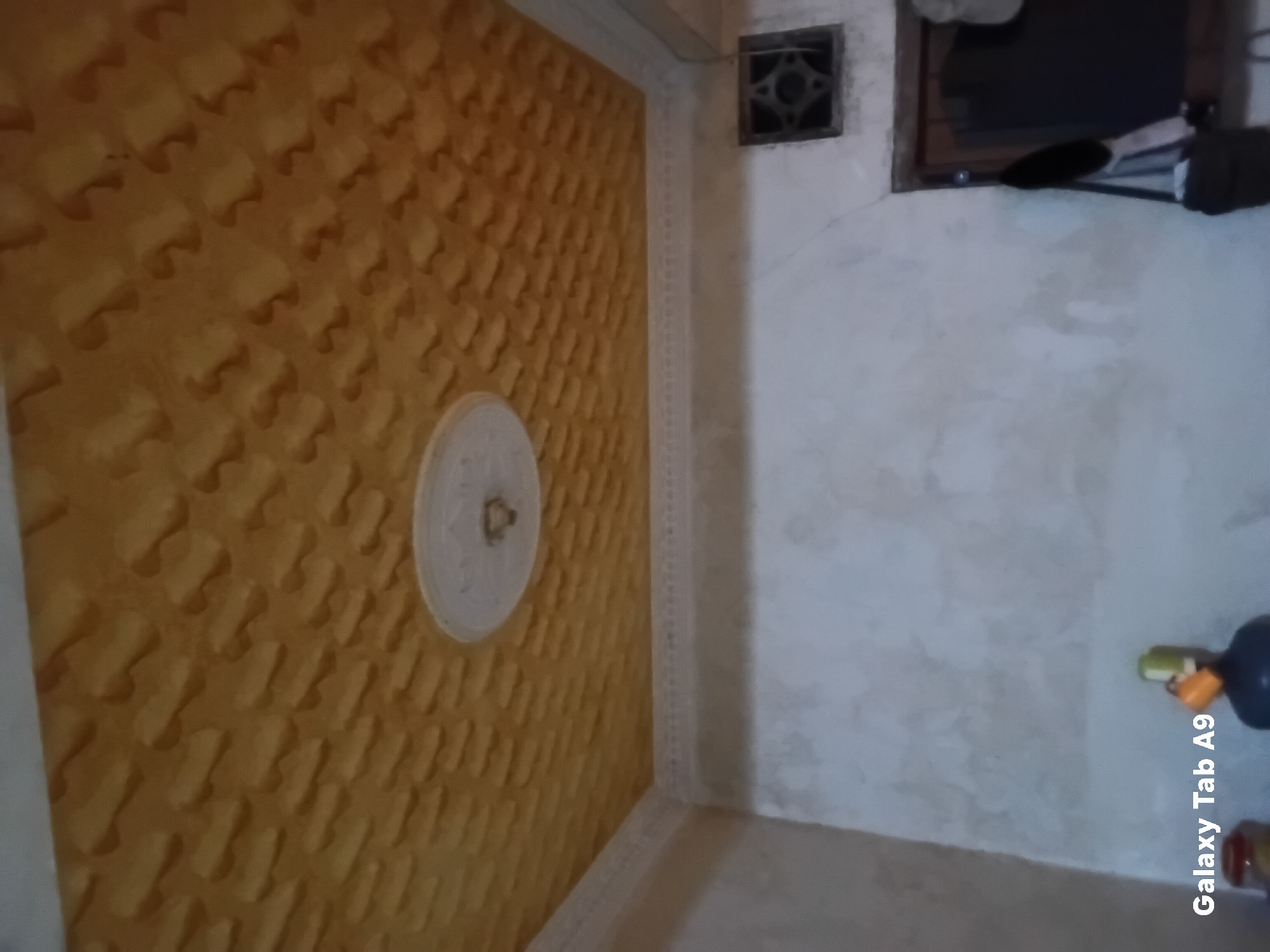 The image shows an interior view of a room with a textured yellow ceiling and a white circular fixture in the center. The walls are plain and light-colored. In the lower part of the image, there is a partial view of a doorway with a dark door and a small, dark square vent above it. To the left of the door, there is a bag. On the far left of the image, the text "Galaxy Tab A9" is visible, suggesting this image was taken with a Samsung Galaxy Tab A9 device. No people are visible in the scene. The environment appears to be a residential or simple commercial space. Without additional information or context, it is not possible to determine the specific location within the city of Trenggalek, Indonesia.