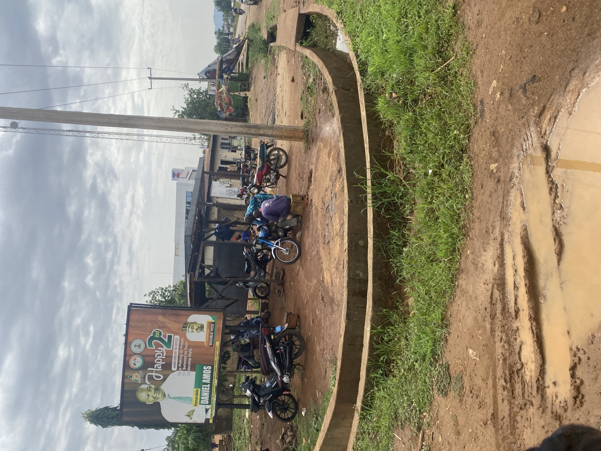 The image, when viewed upright, presents an outdoor scene in Maigizo, Nigeria, featuring a bustling roadside area under an overcast sky. The unpaved ground, particularly on the right side, is muddy with visible puddles and tire tracks, indicating recent rain or poor drainage. A concrete drainage ditch or canal with grassy edges runs along the right side of the frame.
In the foreground and midground on the left, a large political billboard is prominently displayed. It celebrates "Honorable Daniel Amos'" "Happy 2nd Anniversary in Office," praising his "Leadership Style" and identifying him as "Member Representing The Good People of Jema'a/Sanga Federal Constituency." The billboard also features a political party logo, likely the All Progressives Congress (APC), and the text "Courtesy: DOUBLE VICE."
Beneath the billboard and further into the midground, several people are gathered, some sitting or standing near numerous parked motorcycles. One individual in a purple shirt is hunched over something, while another wears a blue patterned outfit. These individuals appear to be engaged in daily activities, possibly at a roadside stall or gathering point. In the background, simple, low-rise buildings with corrugated metal roofs and awnings are visible, along with sparse trees and utility poles traversing the cloudy sky. The scene captures a slice of everyday life in a semi-urban or rural Nigerian setting, characterized by local commerce, political advertising, and basic infrastructure.