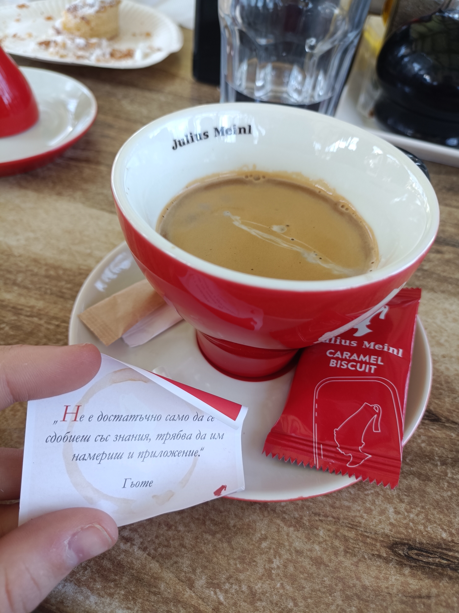 A close-up indoor shot depicts a red and white Julius Meinl coffee cup on a matching saucer, a red Julius Meinl Caramel Biscuit wrapper, and a small card with a quote in Bulgarian. The coffee cup is filled with a light brown liquid, likely coffee. A sugar packet is also on the saucer. In the foreground, a hand holds a card that reads, "Не е достатъчно само да се сдобиеш със знания, трябва да им намериш и приложение." and is attributed to Гьоте (Goethe). A partial plate with food remains and a glass can be seen in the background. The scene is set on a light brown wooden table. This is a common sight in a cafe, likely in Plovdiv, Bulgaria, where Julius Meinl coffee is popular.