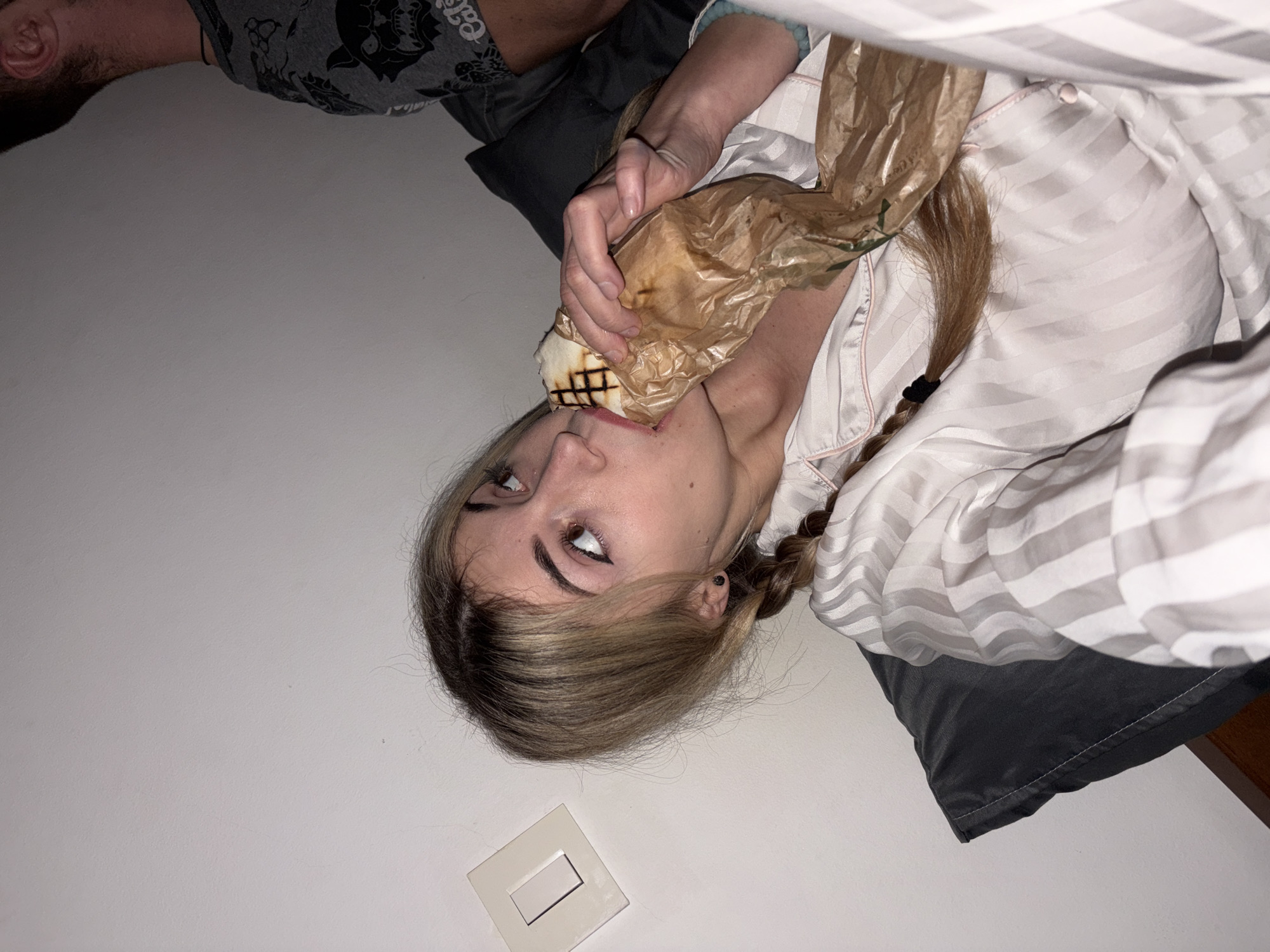 The image depicts an indoor scene featuring two individuals. A young adult female, positioned in the foreground and center-left, is seen reclining against a dark gray pillow. She has light-colored hair with darker roots, styled in two visible braids on either side of her head. Her left eye, with a prominent winged eyeliner, is visible, and she is looking upwards and to her right. She wears light-colored, possibly white or cream, striped pajamas with pink piping. Her hands are holding a partially consumed food item, appearing to be a grilled flatbread or wrap with distinct grill marks, encased in crinkled brown paper, which she is biting into.
To the upper right of the frame, the right ear, neck, and shoulder of a male are partially visible. He is wearing a dark gray short-sleeved t-shirt with black graphic patterns, including what appears to be a stylized animal head (possibly a cat or skull) and partially visible text that reads "Cals".
The background consists of a plain white or off-white wall. A light switch with a rectangular toggle is mounted on this wall to the left of the female's head. No specific architectural details, landmarks, or unique identifiers are present in the image to confirm the location as Budva, Montenegro. The setting appears to be a casual, private indoor space, possibly a bedroom or living area.