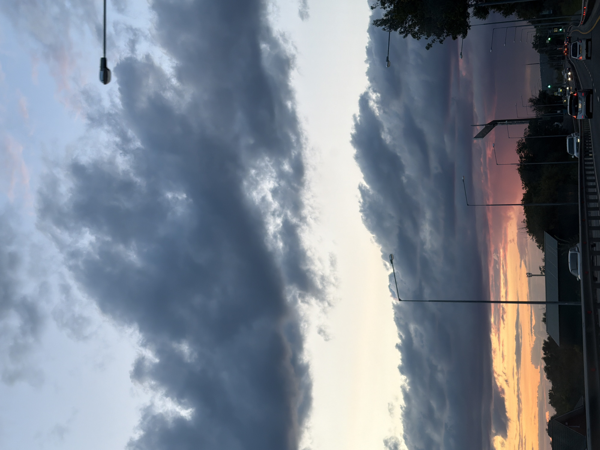 The image captures a twilight scene on a highway, likely in Samara, Russia, given the context. The sky is dramatic, with heavy, dark clouds dominating the upper portion, transitioning to softer hues of pink and orange near the horizon. The setting sun is visible as a vibrant glow behind the clouds, casting a warm light.
The highway itself is multi-lane, with traffic visible. Several cars are driving, with headlights and taillights illuminated, indicating it is dusk or early evening. Streetlights line the side of the road, some of them already on. A protective barrier separates the highway from the area alongside it.
On the sides of the highway, there are trees and vegetation, typical of an urban or suburban landscape. In the foreground, parts of a road barrier are visible. There are no people clearly discernible in the image, but the presence of cars and streetlights suggests human activity and infrastructure. The overall impression is one of a journey on a highway as day turns into night, with the dramatic weather adding a sense of atmosphere.