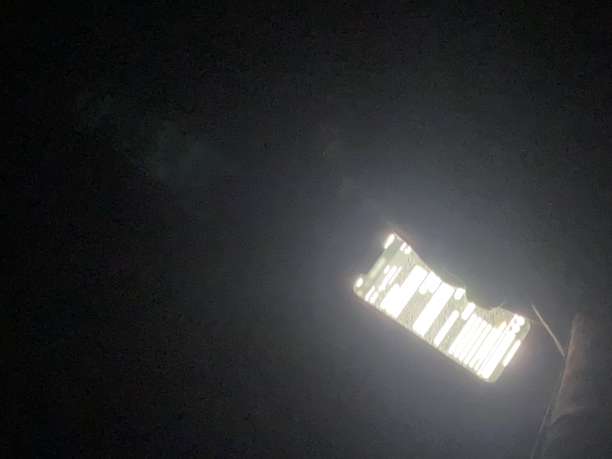 A cell phone screen is brightly lit against an otherwise dark background. The phone screen displays a grid-like pattern of white bars. The setting appears to be outdoors at night. The location is undetermined.