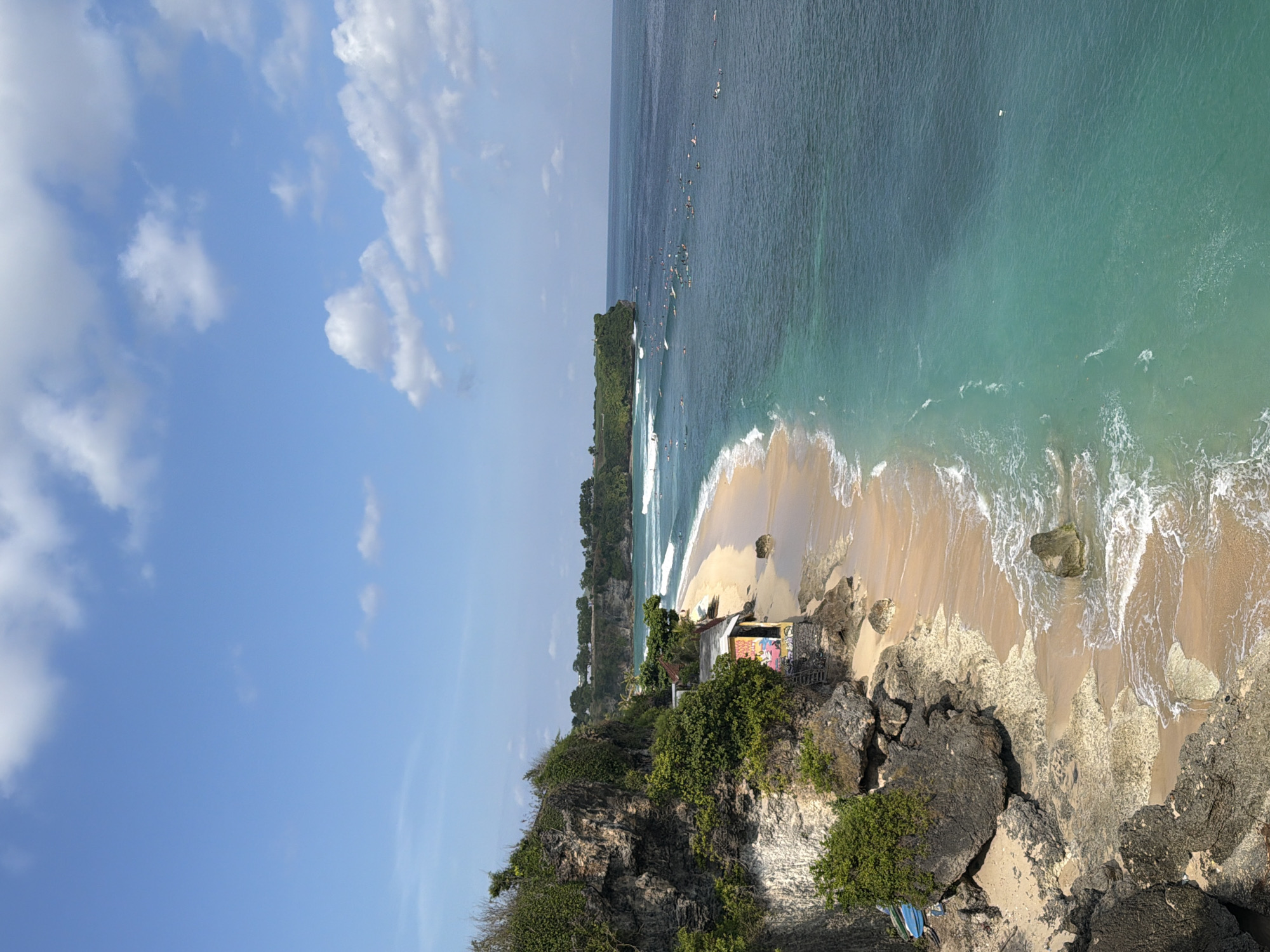 The image displays a high-angle view of a picturesque coastal landscape in Kuta Selatan, Indonesia, under a bright blue sky.
In the foreground, a rugged, rocky cliff face covered with dense green tropical vegetation slopes downwards. On an outcrop of this cliff, overlooking the beach, stands a small, rustic building or shack, its walls adorned with colorful graffiti or painted art. Near the base of the cliff, some blue tarps or fabrics are visible.
A crescent-shaped golden sandy beach unfurls from the cliff's edge into the mid-ground. Along the shoreline, white-crested waves are seen breaking gently, indicating a vibrant surf.
The ocean stretches beyond the beach, exhibiting a beautiful gradient from clear, lighter turquoise near the shore to a deeper blue further out. Numerous small figures on surfboards, appearing as white specks, are scattered across the water, indicating a popular surfing activity underway. A verdant, tree-covered headland juts into the ocean in the mid-distance on the left side, adding to the tropical scenery.
The upper portion of the image is dominated by a clear, bright blue sky dotted with fluffy white clouds. The strong, direct sunlight suggests it is daytime, likely morning or early afternoon, with clear weather conditions. The scene evokes a sense of vibrant outdoor activity and natural beauty, characteristic of a tropical beach destination. No legible text is clearly discernible on any objects.