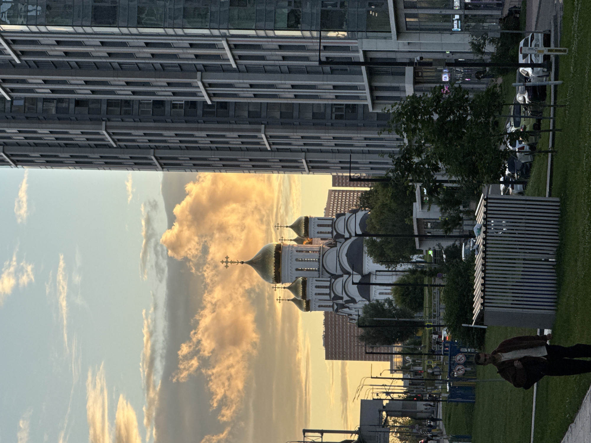 This media file is a photograph taken in Moscow, Russia. The image is oriented vertically, with the top of the frame appearing on the left.
The scene features a striking contrast between modern architecture and a historic religious building. On the left side, a large, contemporary building with a glass facade dominates the upper portion of the frame. Its repetitive windows and horizontal lines suggest a commercial or residential structure. To the right of this modern building, a cluster of white domed churches with golden onion domes stands out. These churches, likely historic, are adorned with crosses on their spires.
The sky is filled with dramatic clouds, illuminated by the warm light of either sunrise or sunset, casting a golden hue. This suggests the photograph was taken during either the early morning or late evening.
In the foreground, on the right side of the image, a person is walking on a grassy area next to a pathway. The individual is wearing a dark jacket and pants. Further to the right, a striped metal fence runs along the grassy area. Beyond the churches, there are glimpses of more urban elements, including traffic lights, street signs, and other buildings, indicating the scene is in an urban environment. A few cars are also visible on a road in the background.
There is some visible text on a street sign which reads "ОХОТНЫЙ РЯД" (Okhotny Ryad) in Cyrillic. The overall mood is serene, with the warm lighting and the juxtaposition of old and new creating an interesting visual narrative.