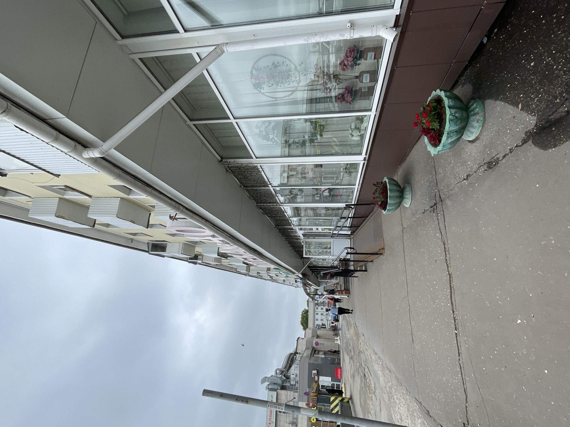 The image depicts a street-level view of a commercial building in Kirov, Russia, under an overcast sky. The building, painted in pale yellow, features a row of large windows, some adorned with floral arrangements visible through the glass. A white awning extends over the storefronts, with a white gutter system running along its edge. Two decorative planters with red flowers are positioned on the sidewalk in front of the building. The sidewalk itself is made of concrete, showing signs of wear and a prominent crack running down its length. Several individuals are walking along the sidewalk, some entering or exiting the stores. In the background, other buildings and infrastructure, including ventilation systems, are visible. A pole with a sign bearing Cyrillic text stands on the left side of the frame. A small bird is captured in flight against the sky.