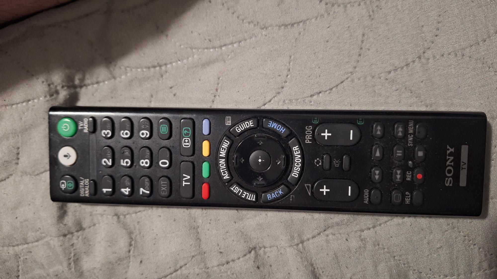 A black Sony television remote control is prominently displayed, lying horizontally on a light-colored, textured fabric surface, likely a blanket or bedspread. The fabric features a quilted pattern of stitched lines. The remote is rectangular with rounded corners and features a multitude of buttons, indicating various functions.
Key sections of the remote include a power button (green) and an input/radio button at the top left, followed by a numeric keypad (0-9) with corresponding letters (e.g., "2abc", "8tuv"). Below these are "DIGITAL/ANALOG" and "EXIT" buttons.
Towards the center, a circular navigation pad is surrounded by function buttons labeled "ACTION MENU", "GUIDE", "HOME", "DISCOVER", "TITLE LIST", and "BACK". Four colored buttons (blue, yellow, green, red) are also visible in this area.
On the right side of the remote, there are buttons for "PROG +" and "PROG -" (channel up/down), volume controls ("+" and "-"), and playback controls including play, pause, stop, fast forward, rewind, skip, and a red "REC" button. Other buttons include "AUDIO", "HELP", and "SYNC MENU". The "SONY" logo is clearly visible on the lower right, with "TV" written vertically next to it. The remote appears to be well-used, with some dust visible on its surface. The overall setting suggests an indoor environment.