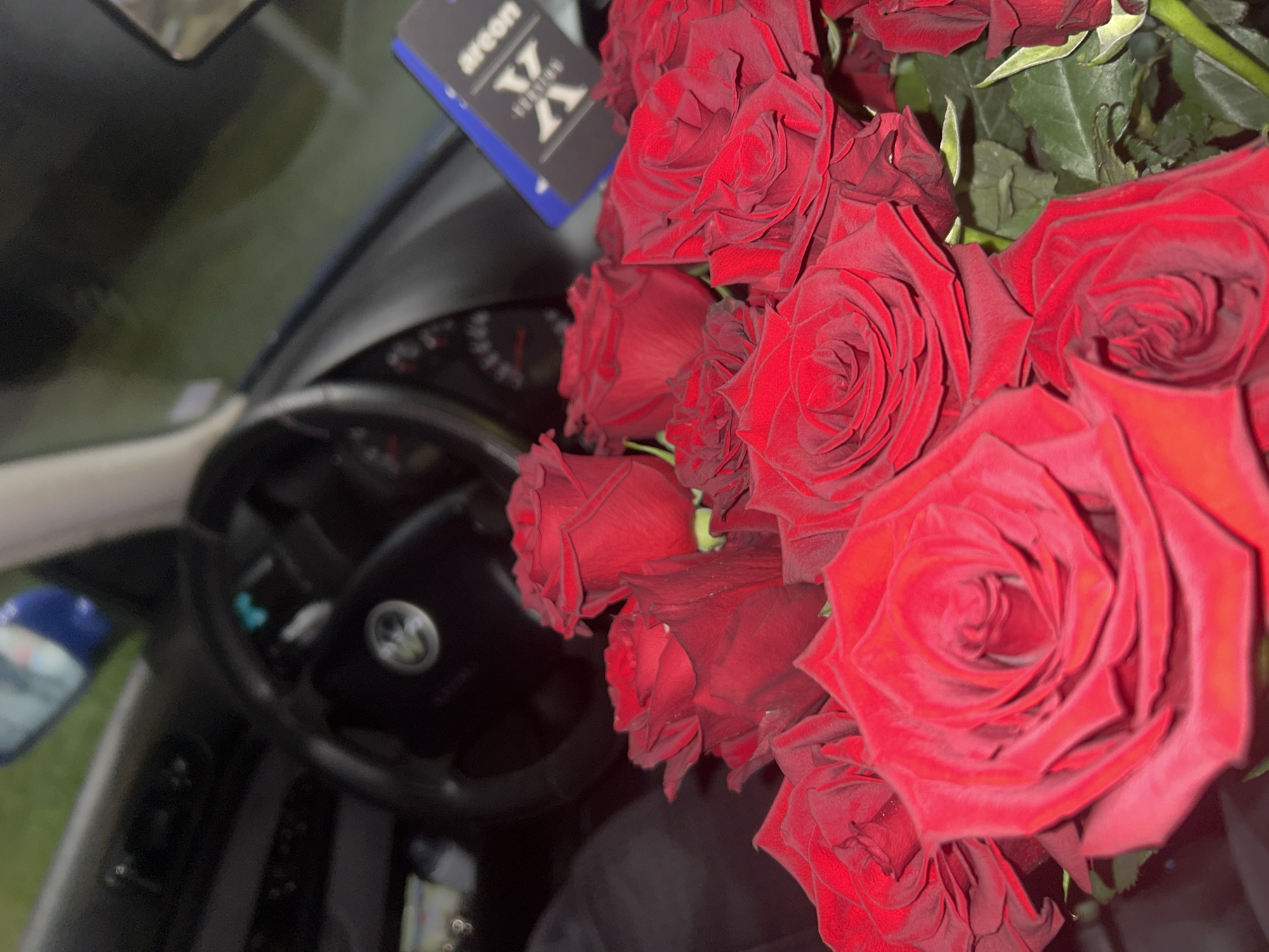 The image shows a bouquet of red roses placed inside a car, likely on the passenger seat or dashboard. The car's steering wheel with a prominent emblem is visible. Also visible is a car air freshener of the brand "Areon". It is not possible to determine the location in Zlatograd, Bulgaria from the given image. No people are visible.