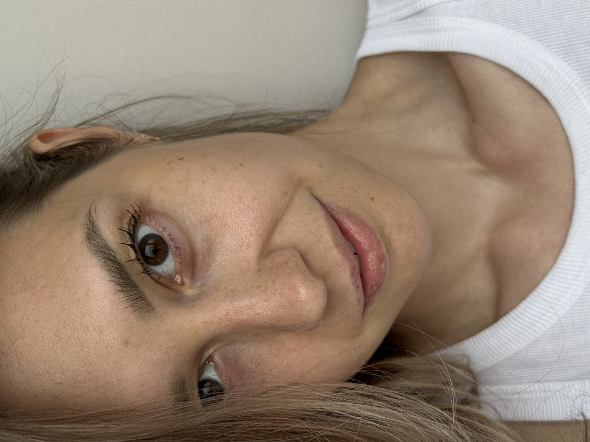 A close-up, slightly rotated image features a woman's face. She has fair skin with freckles and dark eyes. Her hair is light brown and blonde, partially obscuring the lower part of her face. She is wearing a white t-shirt. The background is a plain light color. There is no information to suggest the picture was taken in Chaikovsky, Russia.
