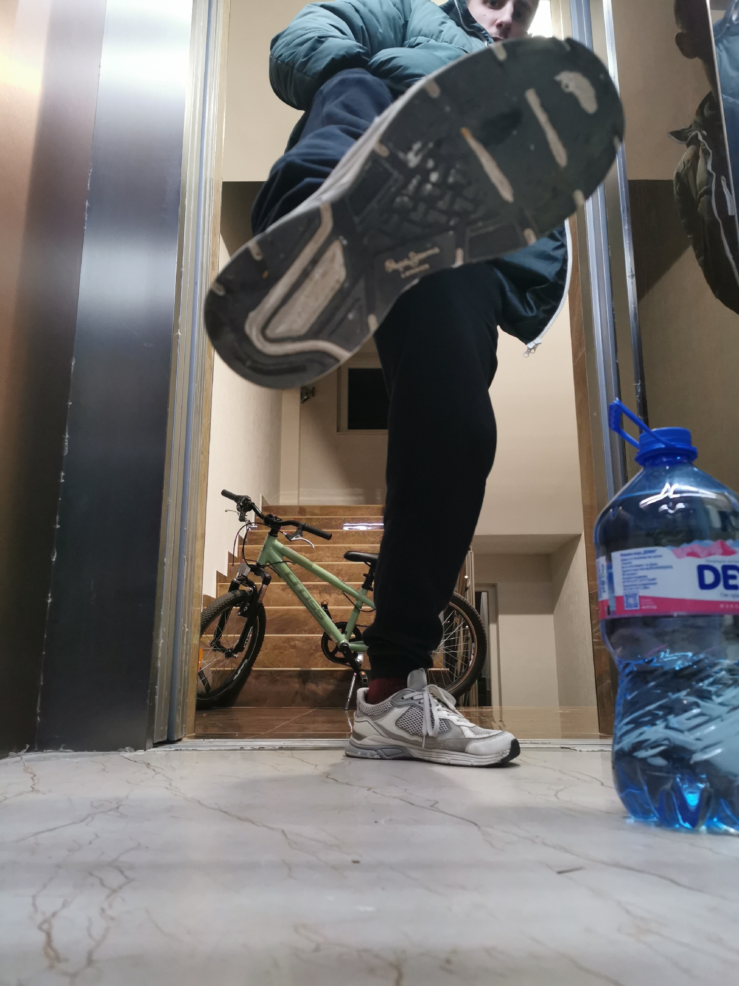 A low-angle indoor photograph captures a dynamic scene, likely in a building's entrance or hallway. In the immediate foreground, a person is shown with one foot raised towards the viewer, prominently displaying the sole of a white and grey sneaker. Faintly visible on the sole is the brand name "Pepe Jeans." The person wears a dark green puffer jacket and black pants, with their other foot visible on the light-colored, marble-patterned floor to the left of a large plastic water bottle. The person's face is partially visible at the top of the frame, looking towards the camera.
To the right in the foreground, a large, clear plastic water bottle with a blue cap and handle sits on the floor. It contains a blue-tinted liquid, and part of its label reads "DE", suggesting a brand like DEVIN, with other unreadable text and QR codes present.
In the mid-ground and background, a light green mountain bike with black tires and handlebars is parked on a set of wooden or tiled stairs that ascend upwards. To the left, a tall, metallic, reflective surface resembling an elevator door or a polished entrance is visible. The walls in the background are plain and light-colored. The overall setting is an urban indoor environment, consistent with a residential or commercial building in Burgas, Bulgaria. The lighting is artificial and evenly distributed.