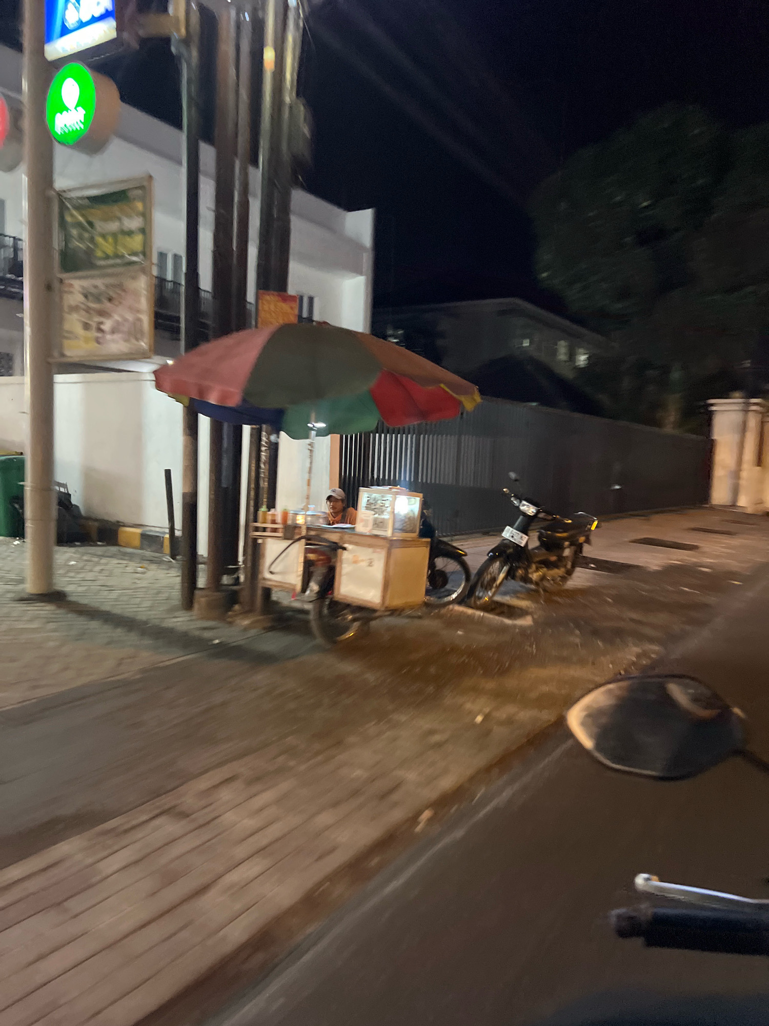 A street food vendor operates a cart at night in Sleman, Indonesia. The cart is under a colorful umbrella, and a person is seated behind it. There are bottles of condiments on the cart. Two motorcycles are parked nearby. The street is paved with bricks, and there are buildings and street lights in the background. The image appears to be taken from a moving vehicle, indicated by the motion blur.