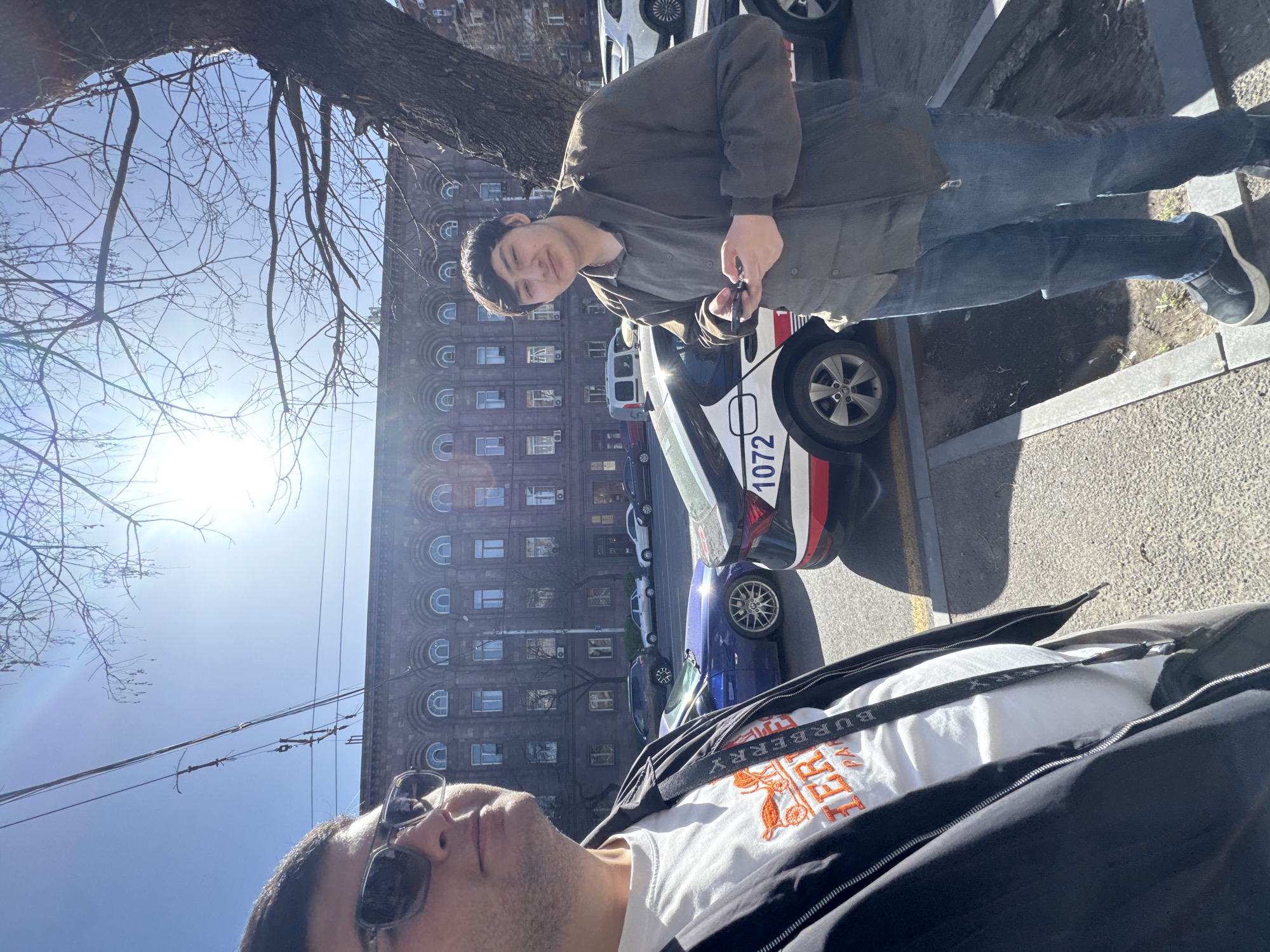 Two men are posing for a picture on a sunny day in Yerevan, Armenia. One man is wearing a brown jacket and jeans and is standing near a parked car. The other man is wearing a white shirt with a Burberry logo and sunglasses. The building in the background is located on Vazgen Sargsyan Street. Several cars are parked along the street. A leafless tree branch is visible in the upper-left corner of the frame.