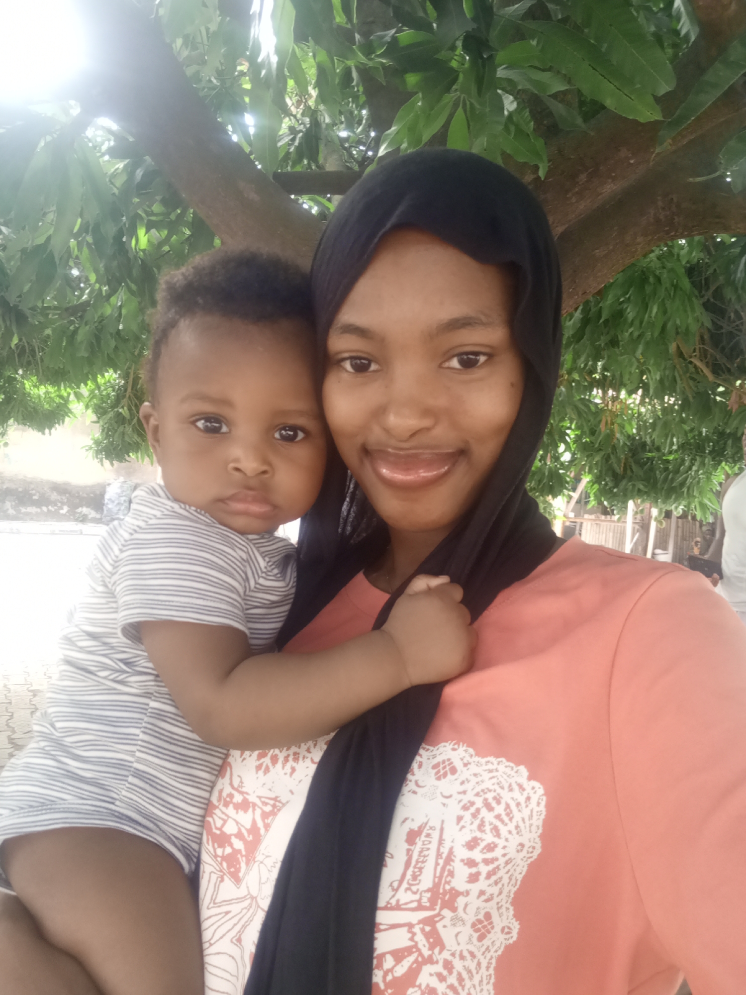 A young woman wearing a black hijab and an orange t-shirt with a white lace-like print on the front is holding a baby. The baby is wearing a white and gray striped shirt. They appear to be outside, under a tree with green leaves. In the background, there is a white wall and a partially visible fence. Based on the prompt context, the location is Keffi, Nigeria.