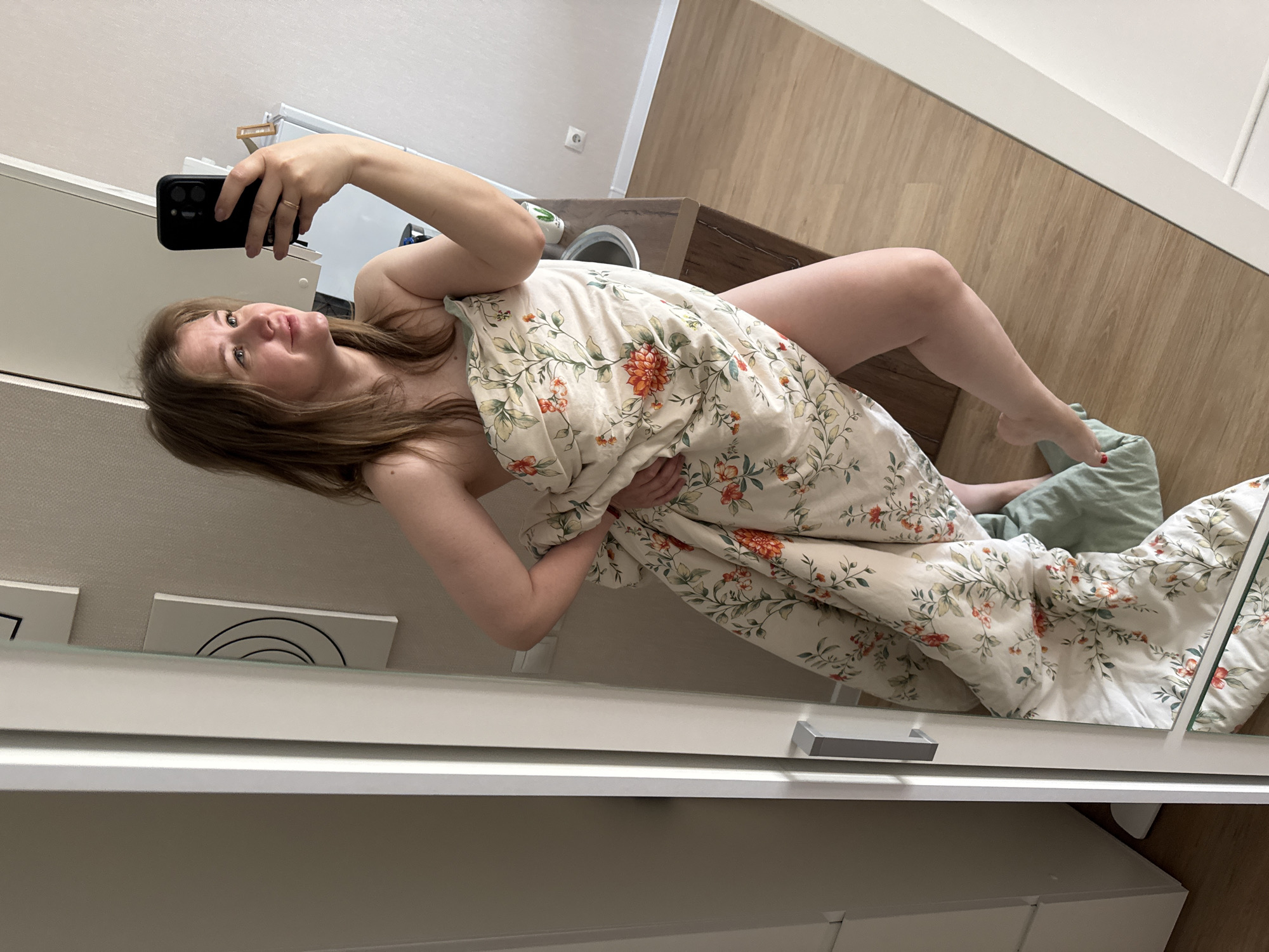The image is a sideways mirror selfie taken indoors, capturing a young woman largely covered by a floral duvet. She is reclining or lying down, holding a black smartphone (resembling an iPhone) to photograph her reflection in a large mirror. Her head is positioned towards the top left of the frame, with her light brown hair spread around her. She looks towards her phone, which is pointed at the mirror. Most of her body is covered by a beige duvet featuring a pattern of green leaves and orange-red flowers. One bare leg and foot are visible towards the bottom right, with red nail polish on her toes, resting on a light green blanket or pillow. The reflected room shows light wooden flooring and white walls. In the reflected background, a white wall outlet is visible above a dark wooden cabinet or counter, next to a white appliance. A white piece of furniture with metal handles is prominent in the mirror's foreground. The scene is an indoor setting, likely a bedroom or dressing area in Perm, Russia. No discernible text is visible within the image.