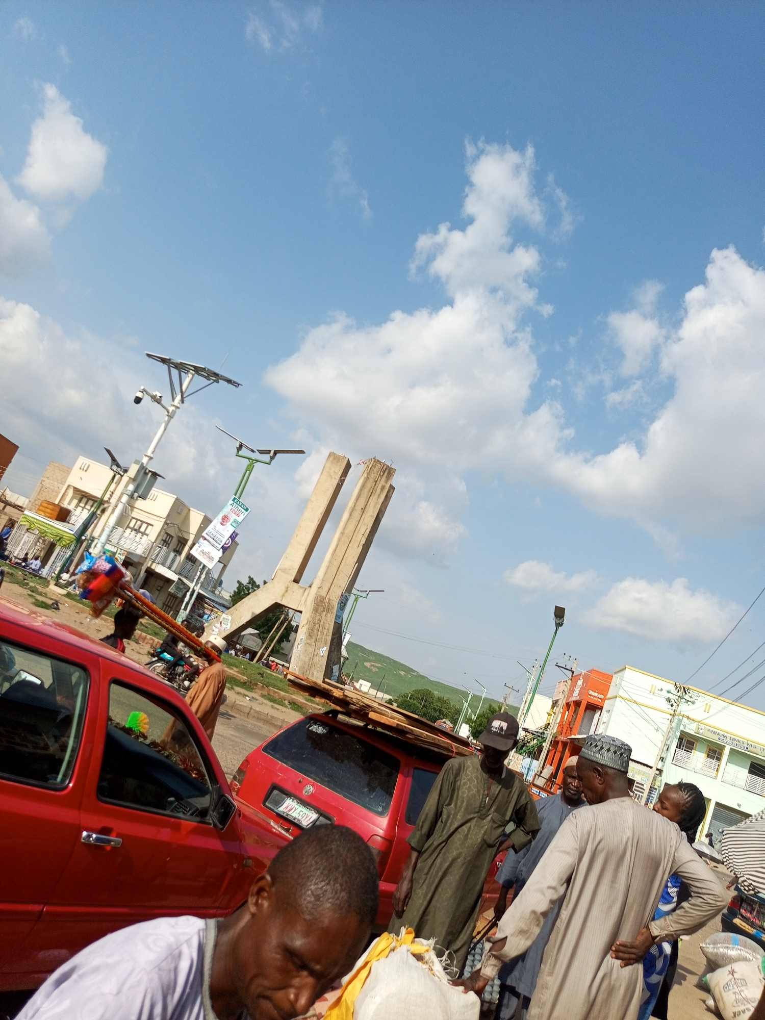 The image shows a street scene in Arawa, Nigeria, with several people and vehicles. In the foreground, a man in a white shirt is partially visible, with a bag of goods in front of him. Behind him, a red car is parked. To the right, another red car is loaded with wooden planks.
Further back, a group of men are gathered, some wearing traditional attire. One man in a green robe appears to be speaking. Other individuals are seen near a building with shops, including one with banners that say "JATA ABUJA 2023".
In the background, there is a concrete structure that appears to be under construction, with tall pillars. Solar-powered streetlights are visible throughout the scene. The sky is bright blue with scattered white clouds. The overall impression is a bustling street market or thoroughfare on a sunny day.