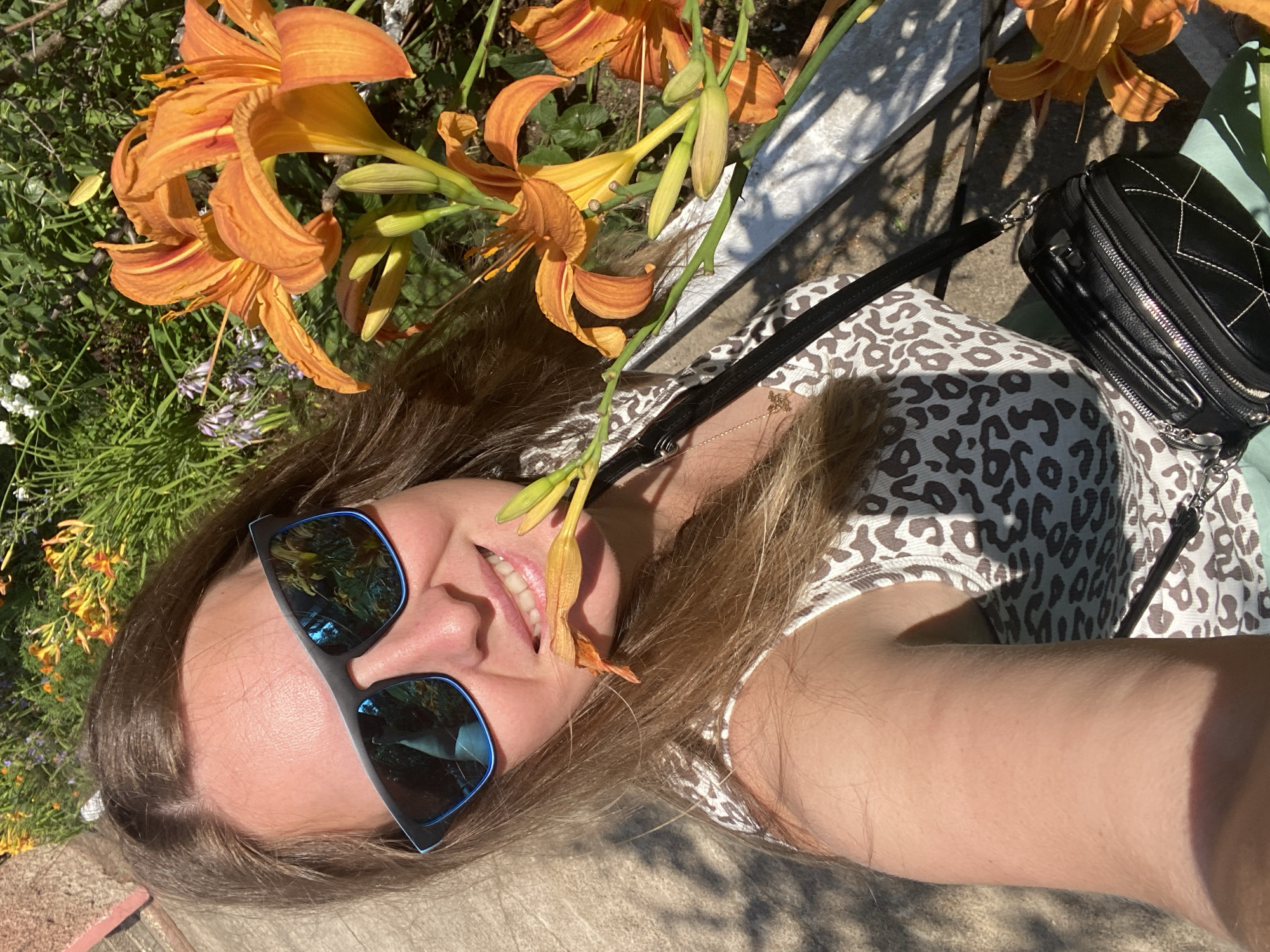 A woman is taking a selfie outdoors, holding a flower to her mouth. She is wearing sunglasses and a leopard-print top. In the background, there are orange lilies and greenery. The scene appears to be a garden or park, possibly in Noviy, Russia, given the foliage. She has a black purse slung over her shoulder.