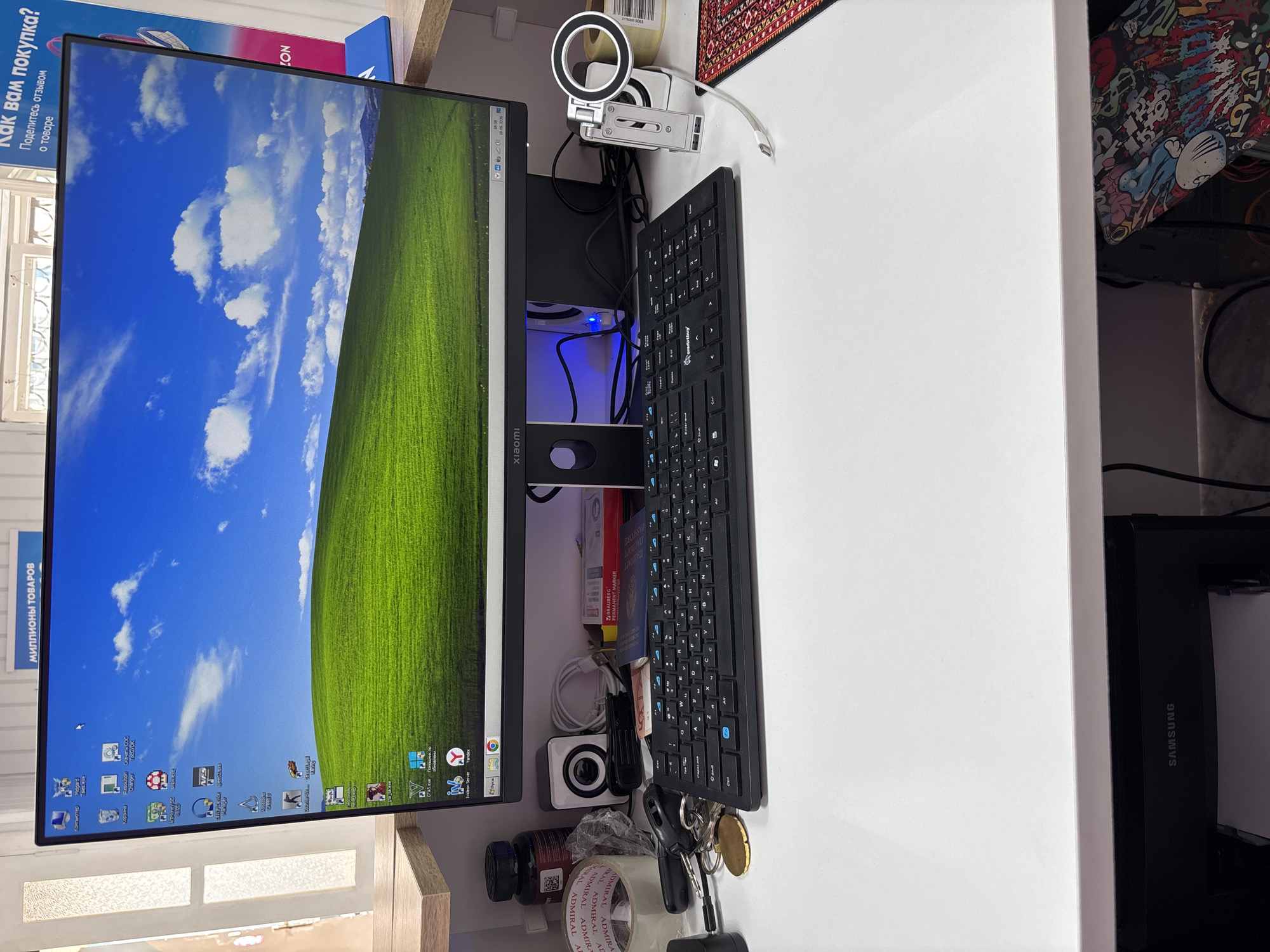 The image depicts a computer desk setup. A large monitor displays the classic "Bliss" wallpaper from Windows XP, showing a green hill under a blue sky with white clouds. On the desk, a black keyboard is positioned in front of the monitor. To the left of the keyboard, there are various items including rolls of tape, a small white speaker, keys with a brass-colored medal, and a dark-colored bottle with a white label. To the right of the keyboard, a USB drive and what appears to be a passport are visible. A mobile phone holder with a ring light attachment is also present on the right side of the desk. In the background, a printer with the "SAMSUNG" logo is visible. The scene is indoors, with a shelf above the desk that has some signage indicating "МИЛЛИОНЫ ТОВАРОВ" (Millions of Goods) and "КАК ВАМ ПОКУПКА?" (How do you like the purchase?). The overall impression is a functional workspace with personal items. There is no indication of the city of Vileyka, Belarus in the image itself.