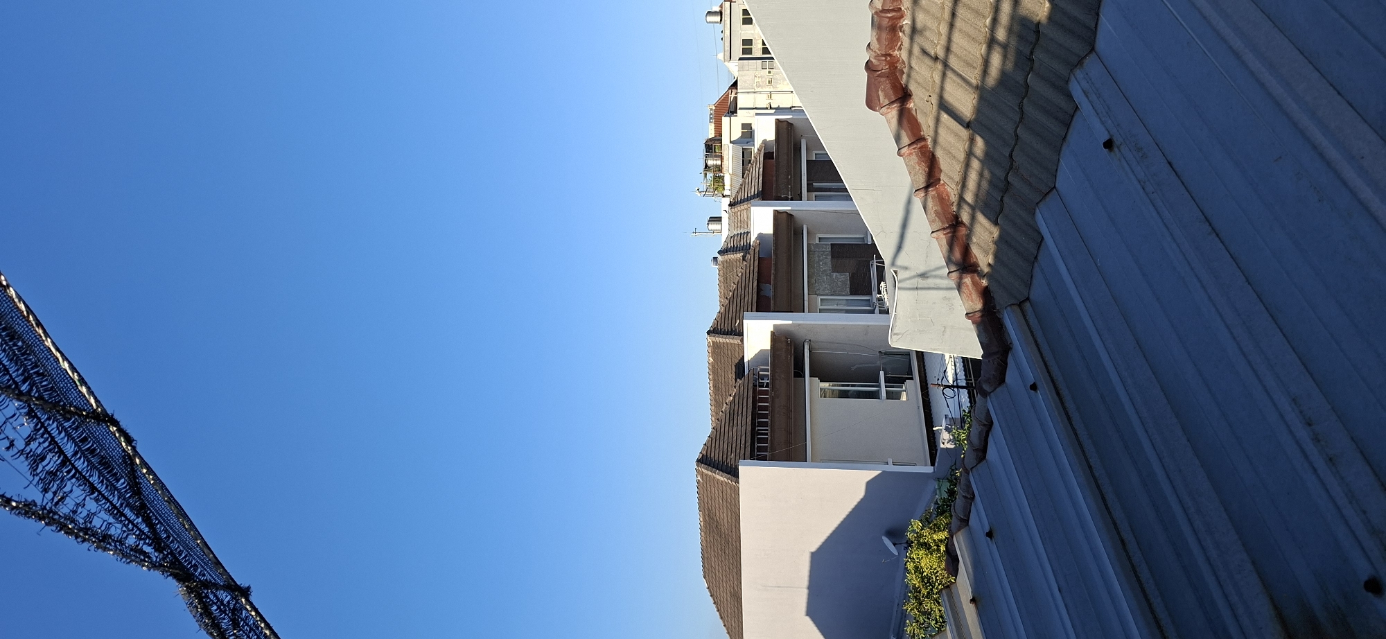 This is a vertical, eye-level, outdoor photograph taken on a clear day with a bright blue sky. In the foreground, a corrugated metal roof takes up the bottom third of the image. Beyond this roof, a second roof made of reddish-brown tiles is visible, partially casting a shadow on the metal roof. In the mid-ground, a row of modern houses with light-colored facades and brown tile roofs are situated. These houses appear to be of a uniform design, with large windows and visible balconies. Some greenery is present on the left side of the houses. The background consists of more buildings and the clear blue sky. The overall scene suggests a residential area in a city, possibly in Surabaya, Indonesia, given the architectural style and the presence of water tanks on some of the rooftops. There are no people visible in the image.