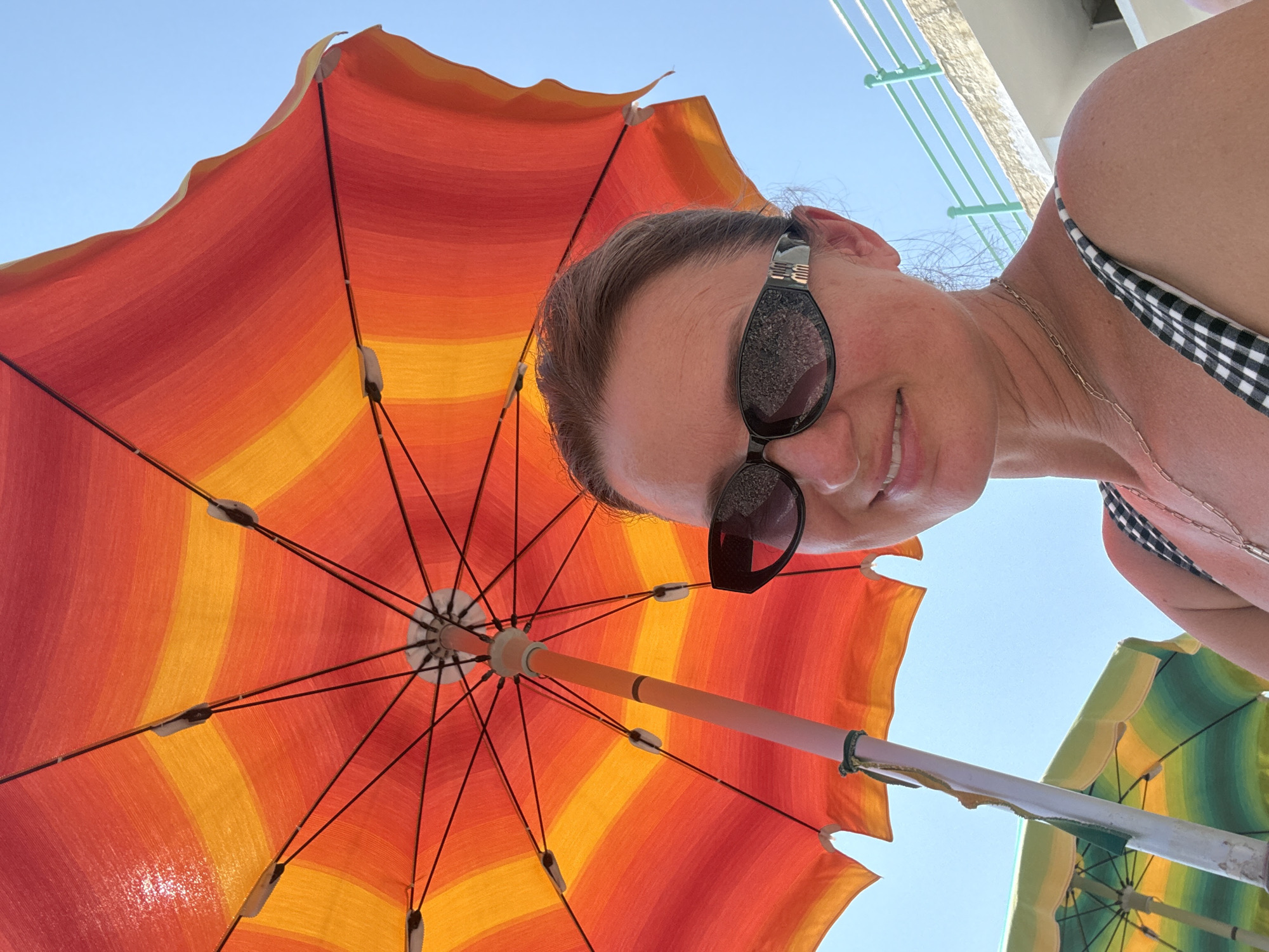 A low-angle selfie shows a woman wearing sunglasses and a black and white checkered bikini top, smiling under a large, orange and yellow striped umbrella. The umbrella's spokes are visible, fanning out from the center. Behind her, another umbrella with green and yellow stripes can be seen. The sky is clear and blue, suggesting a sunny day. The scene appears to be a beach or outdoor recreational area. The woman's sunglasses reflect the surroundings, and she is wearing a thin silver necklace. The image captures a moment of leisure and enjoyment during what seems to be a warm, sunny day, likely in a coastal setting given the umbrellas and beachwear. The time of day is likely daytime due to the bright sunlight. There is no visible text in the image.