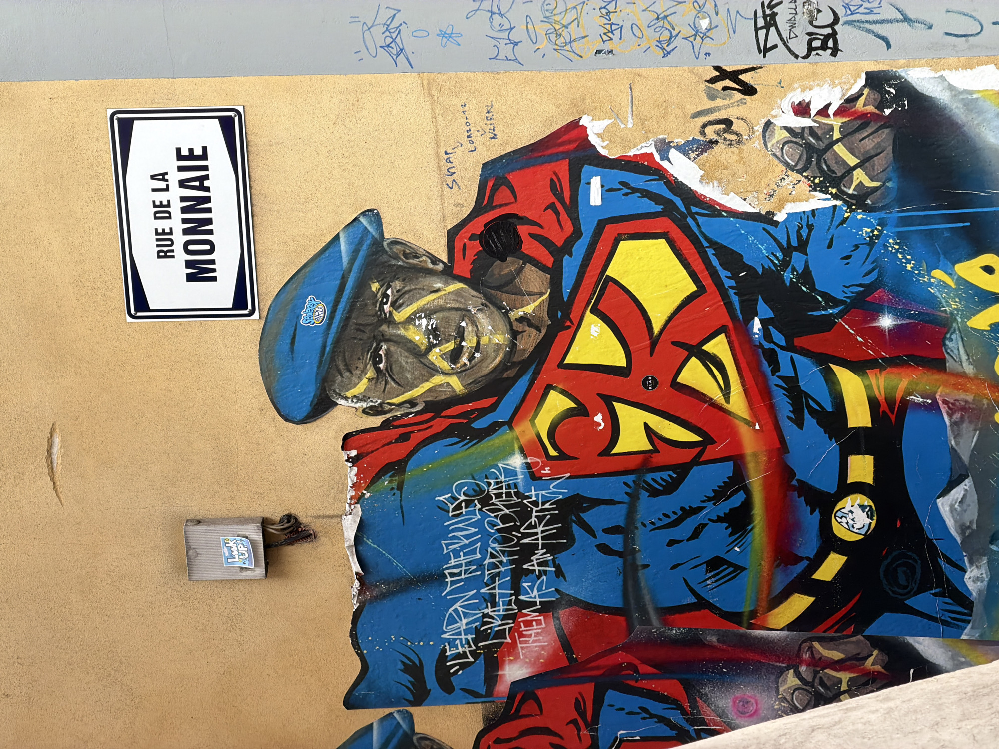 The image depicts a section of a building's exterior wall with graffiti art. A prominent mural features a man dressed as Superman, wearing a blue beret. The man's face is detailed with yellow paint markings. The graffiti is partially damaged and peeling. Above the mural, a street sign reads "RUE DE LA MONNAIE". To the left of the mural, there is a small grey electrical box with a sticker that says "Look UP!". The wall's surface is textured and painted a light orange-yellow color. To the right of the mural, there is additional graffiti on a lighter grey section of the wall. The overall scene appears to be an urban streetscape.