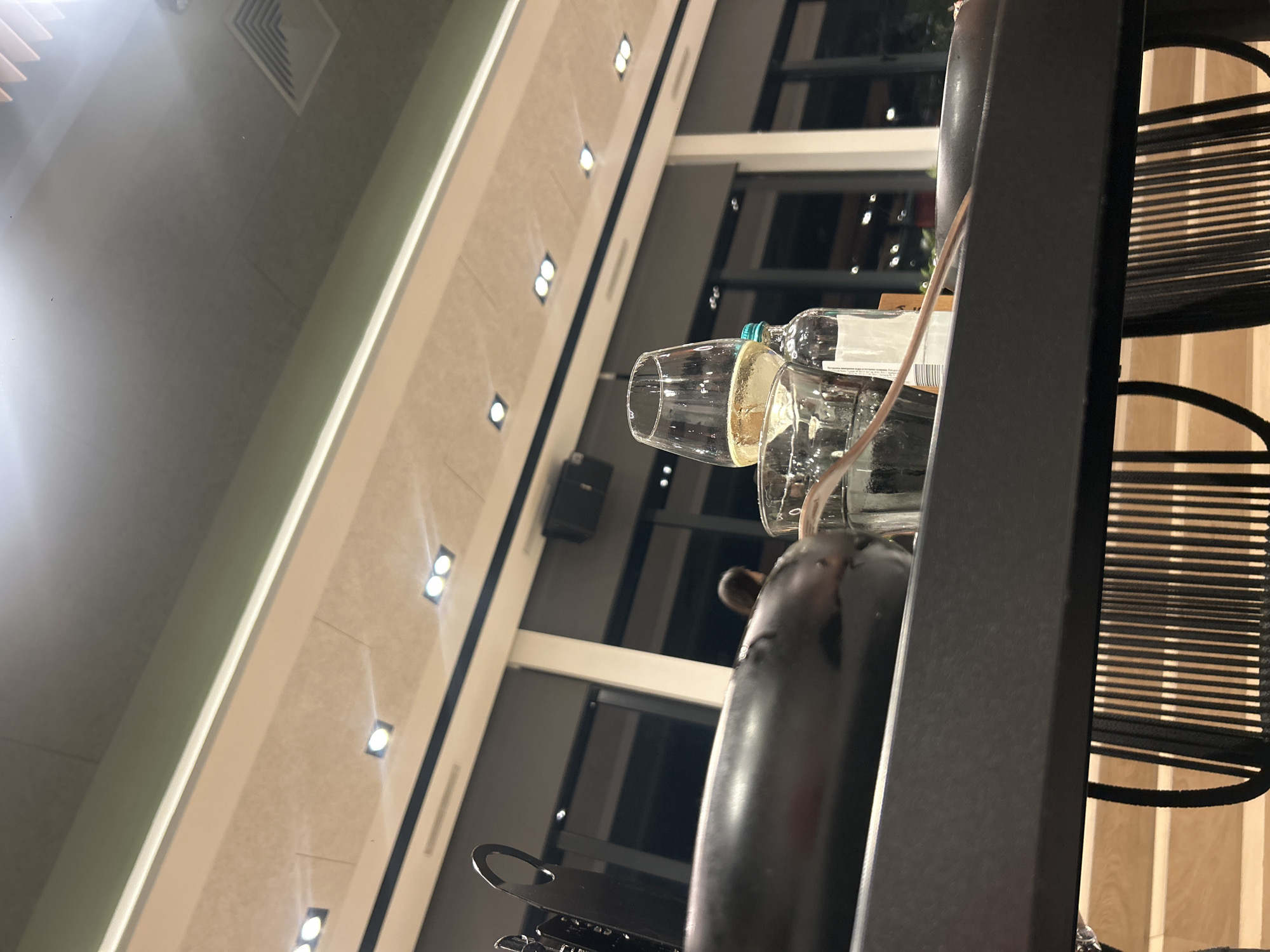 The image is taken indoors, likely in a restaurant or bar. The setting is Plovdiv, Bulgaria. A table setting includes two glasses, one containing white wine. Behind the glasses is a bottle of wine, partially obscured. The focus is on the foreground objects, with a bar frame and chairs visible to the right. The background reveals large windows offering a glimpse of the city’s modern architecture, characterized by clean lines and lights.
