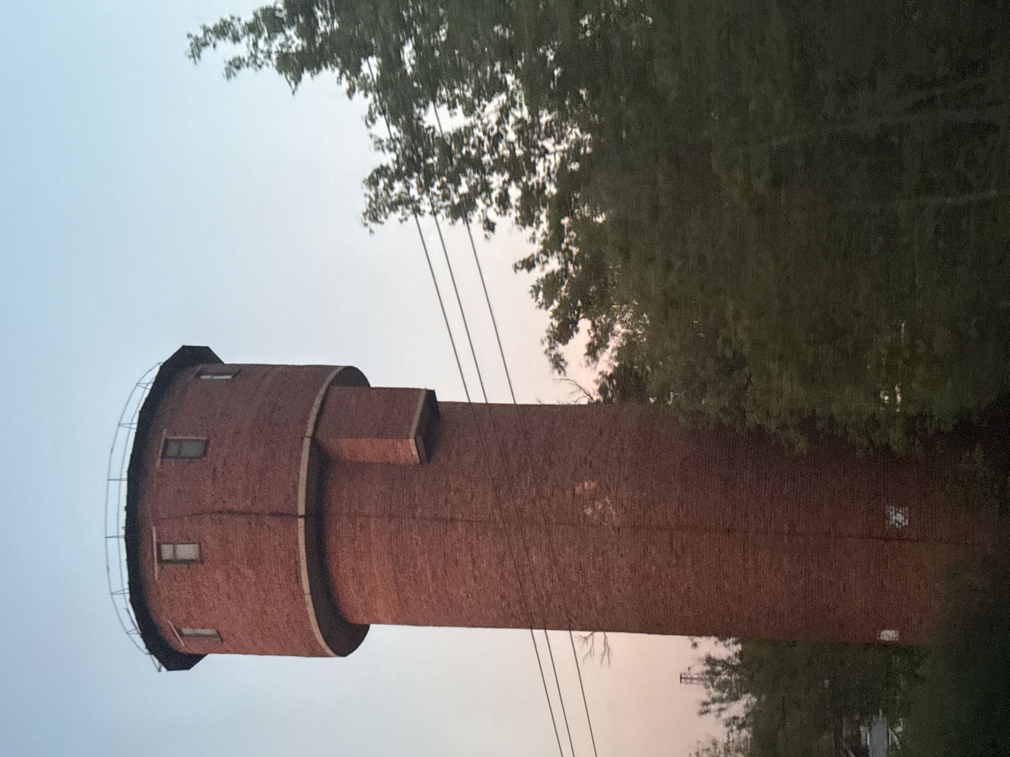 A tall, cylindrical brick water tower stands in Iksha, Russia. The tower is the primary subject, positioned vertically in the frame. It features a smaller cylindrical section near the top, a rectangular protrusion, and several small windows near the top section. The tower's brick texture is visible throughout. Trees and foliage are present on the right side of the image, partially obscuring the tower. Power lines extend from the tower toward the right. The sky is visible in the background, transitioning from light blue to a softer, warmer hue near the horizon.