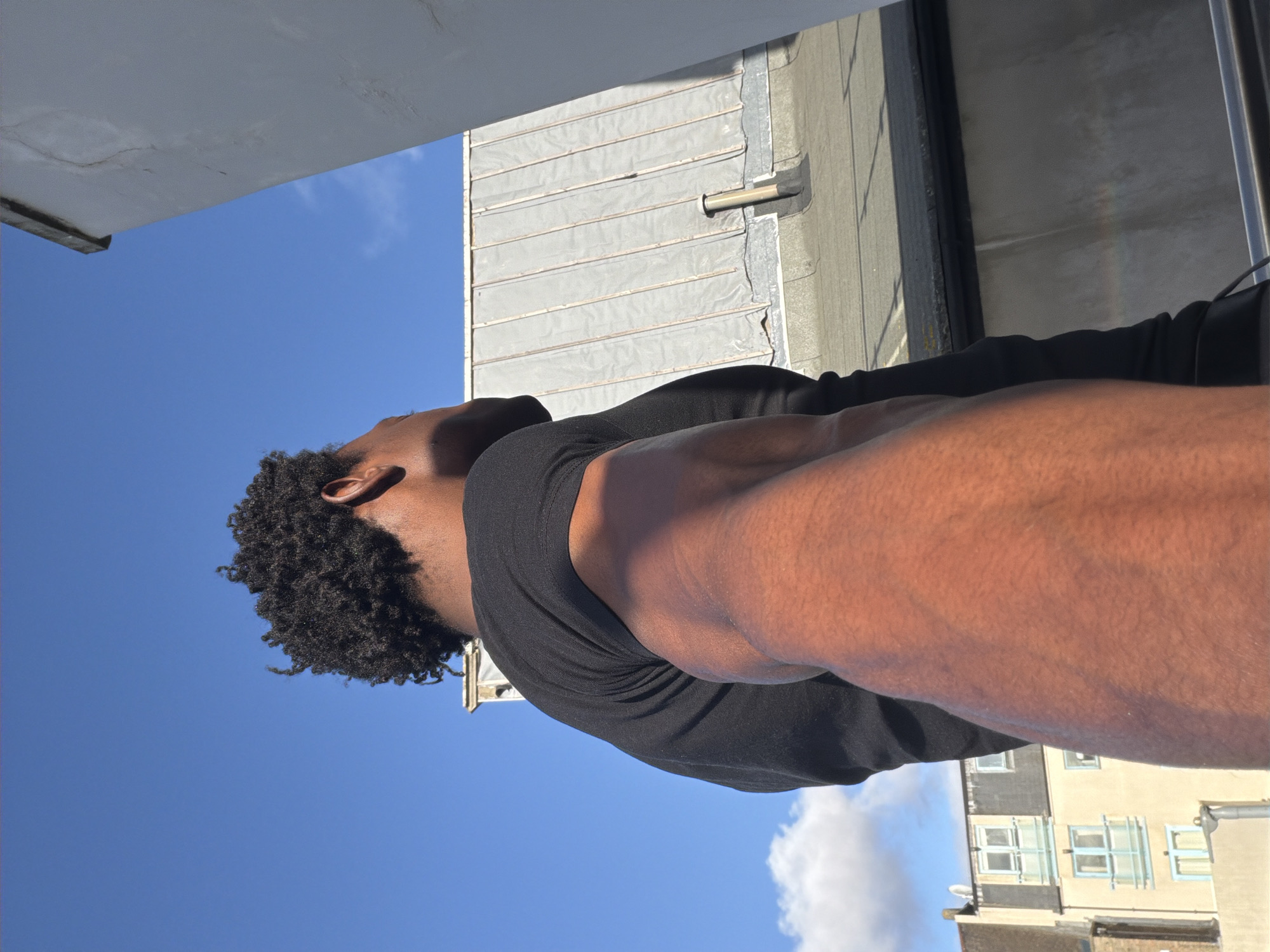 A low-angle shot captures a person from approximately shoulder-level and higher, set against an urban backdrop. The individual, who appears to be male, has dark skin and short, curly black hair. He is wearing a dark-colored shirt or sweater.
The background features architectural elements of a city: a gray, corrugated metal roof of a building, partial views of adjacent buildings, and a clear blue sky with scattered clouds. The architecture includes windows and potentially a satellite dish on one of the buildings.
While specific landmarks cannot be identified, the general architectural style could suggest the image was taken in London, United Kingdom. There is no specific identifiable information to pinpoint the location further within London.