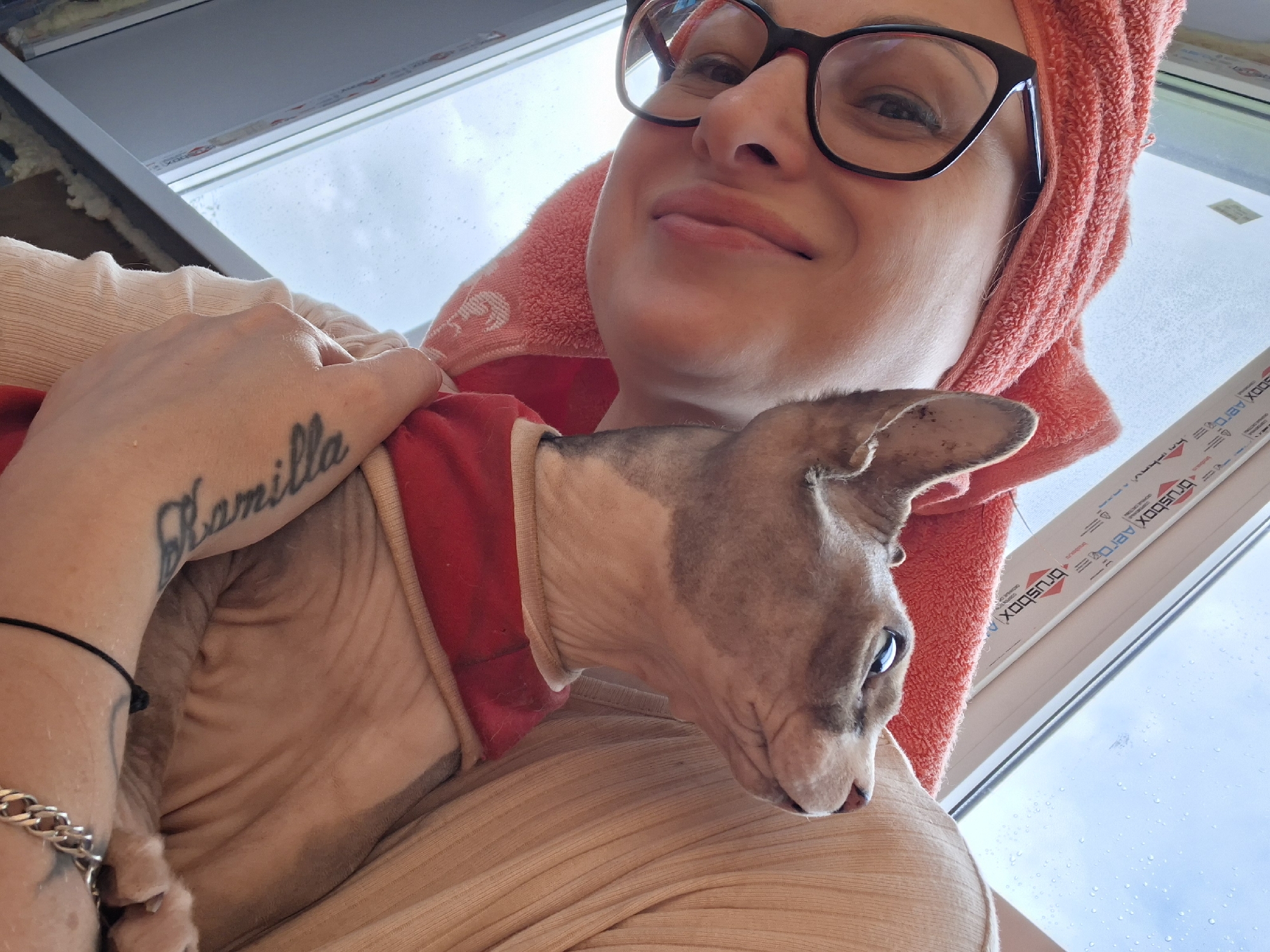 A person wearing glasses and a pink towel on their head is holding a Sphynx cat. The cat is wearing a red garment around its neck. The person has tattoos on their arm. They are positioned near a window with raindrops on the glass, suggesting an outdoor or indoor setting with a view of the weather. The background includes a window frame with some visible text, possibly branding, and insulation material beneath the window. The overall scene appears to be an informal, personal moment captured in what could be a home environment within Забалоцкі сельскі Савет, Belarus.