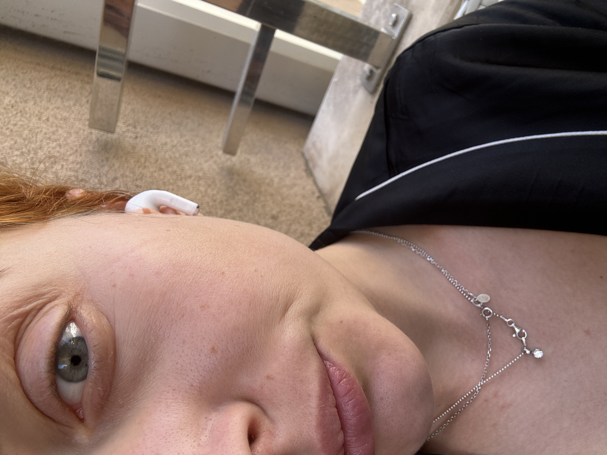 The image is a close-up, low-angle shot of a person lying down, with a partial view of their face, chest, and surrounding environment. The person has fair skin, freckles, and a single visible eye that is grayish-green. There is an earbud in the person's right ear. The person is wearing a black top with white piping and a delicate silver necklace with a small charm.
The background includes a section of carpet and part of a white table or other furniture piece with chrome legs.
There is no information in the image to indicate that this image was taken in the city of Budva, Montenegro.