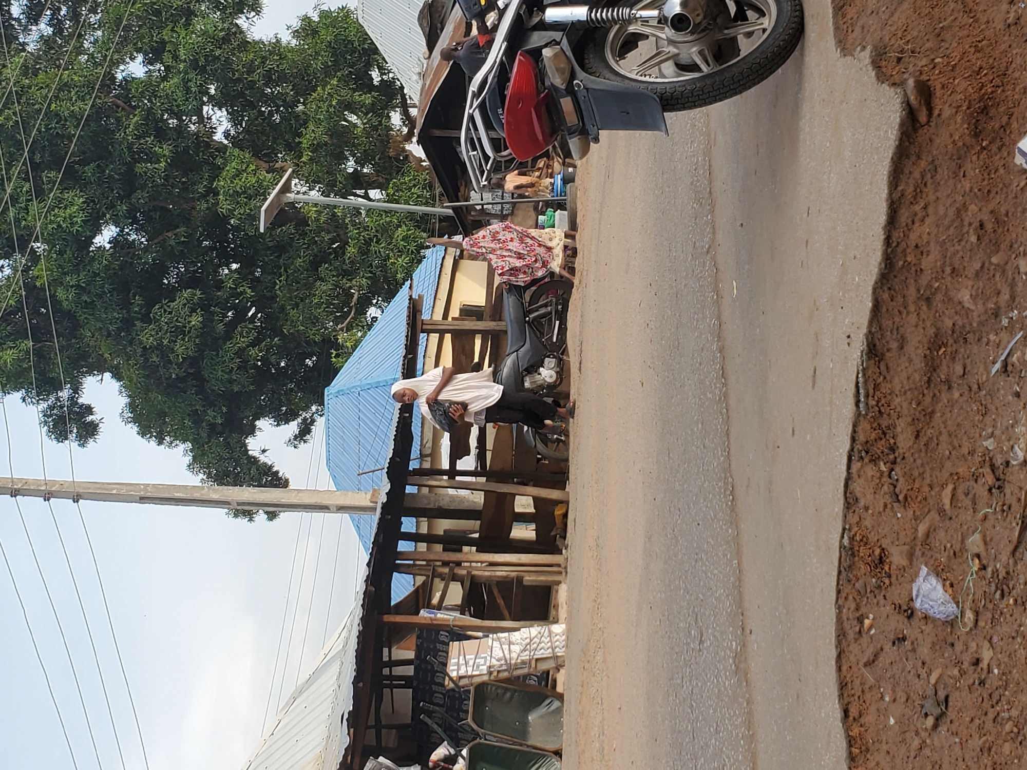 This media file captures a daytime street scene in Mararaba, Nigeria. The image is taken from a slightly elevated perspective, looking down a paved road that runs from the foreground towards the background, flanked by a dirt shoulder on the right.
In the midground on the left, a person is standing next to a dark-colored motorcycle. This individual is wearing a white head covering (hijab), a light-colored long-sleeved top, and dark pants, holding a black bag. Behind them, there are several makeshift stalls or shops constructed with wooden frames and corrugated iron roofs, some with blue-colored sheets. Goods like mattresses and green wheelbarrows are visible within and around these structures. Further back, another person in a patterned dress or shirt (red, white, and black) is partially visible, appearing to be near another motorcycle.
On the far right in the foreground, only the rear section of a different motorcycle is visible, showing a silver wheel, part of the exhaust, and a red taillight. The paved road surface transitions into a dirt area on the right, where some small debris and earth are scattered.
The background on the left is dominated by a large, dense tree with abundant green foliage. Above the stalls and road, power lines stretch across the frame, supported by utility poles. The sky is clear and bright, indicating a sunny day. No discernible text is visible in the image.