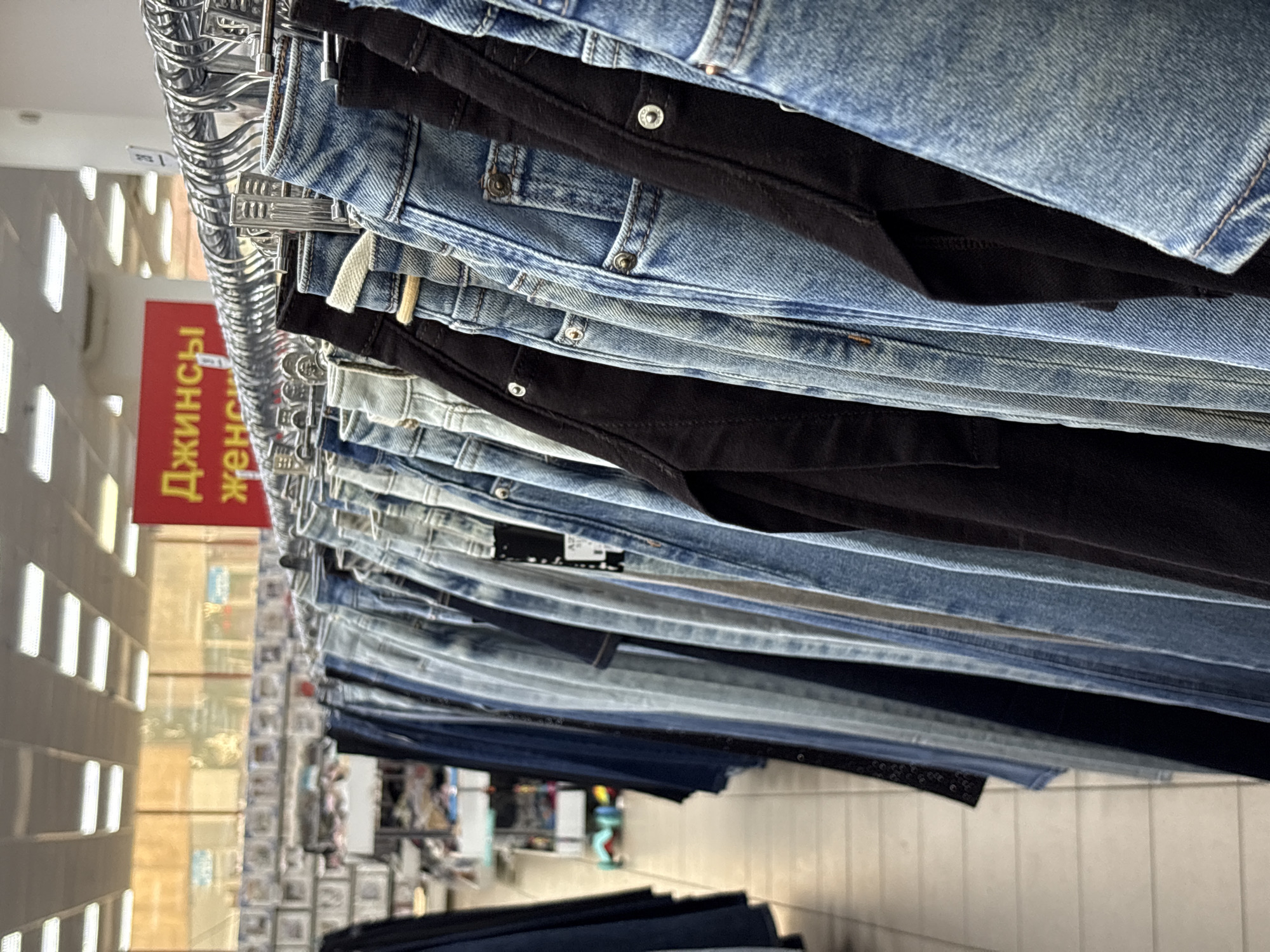 The image depicts a close-up view of a clothing display inside a retail store, likely in Votkinsk, Russia. The primary subjects are numerous pairs of jeans, neatly hung on metal hangers along a rack. The jeans exhibit a range of washes and colors, predominantly various shades of blue denim and black, with some lighter-colored pairs also visible. They are displayed in a stacked manner, with the waistbands and upper portions of the trousers facing the viewer.
The perspective is angled upwards and sideways, providing a detailed look at the texture of the denim and the metal hangers. In the mid-ground, a prominent red sign with white/yellow Cyrillic text is visible. The text reads "Джинсы женск...", which translates from Russian as "Jeans women's...", indicating that this section is dedicated to women's jeans.
The background is somewhat blurred but shows typical elements of a store interior, including shelves or additional merchandise displays. Overhead artificial lighting, visible as bright rectangular fixtures, illuminates the scene. The floor appears to be tiled. No people are present, and no specific activity is taking place other than the organized display of clothing for sale. The scene conveys a sense of an ordinary, well-stocked commercial environment.