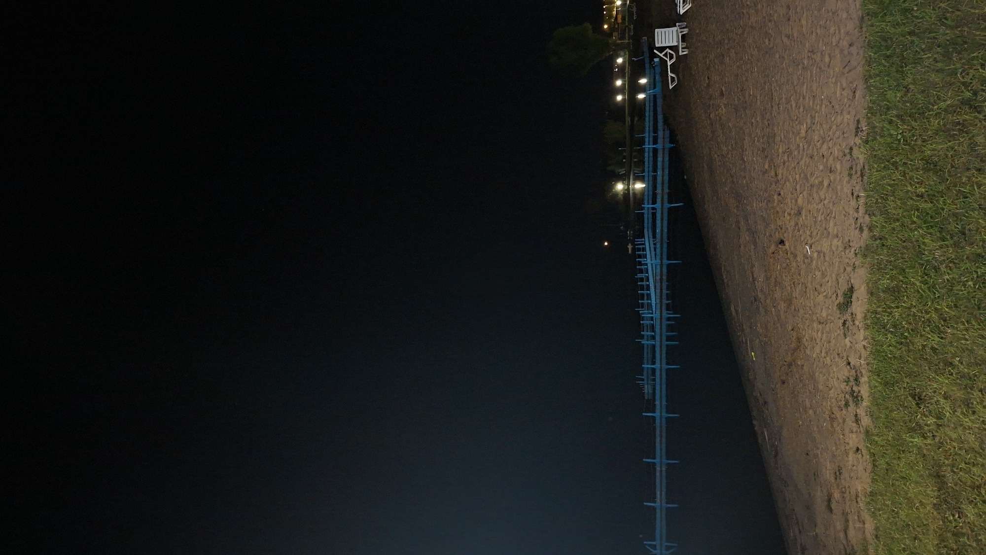 A night-time view of a sandy beach area adjacent to a body of water. A blue, horizontal structure, possibly a pier or a floating barrier, extends across the water from left to right. On the beach, there are two white lounge chairs. In the background, a line of lights is visible, suggesting a walkway or buildings along the shoreline. The sky is dark and clear. The scene appears to be a public recreational area. The location is identified as Myadzyel, Belarus.