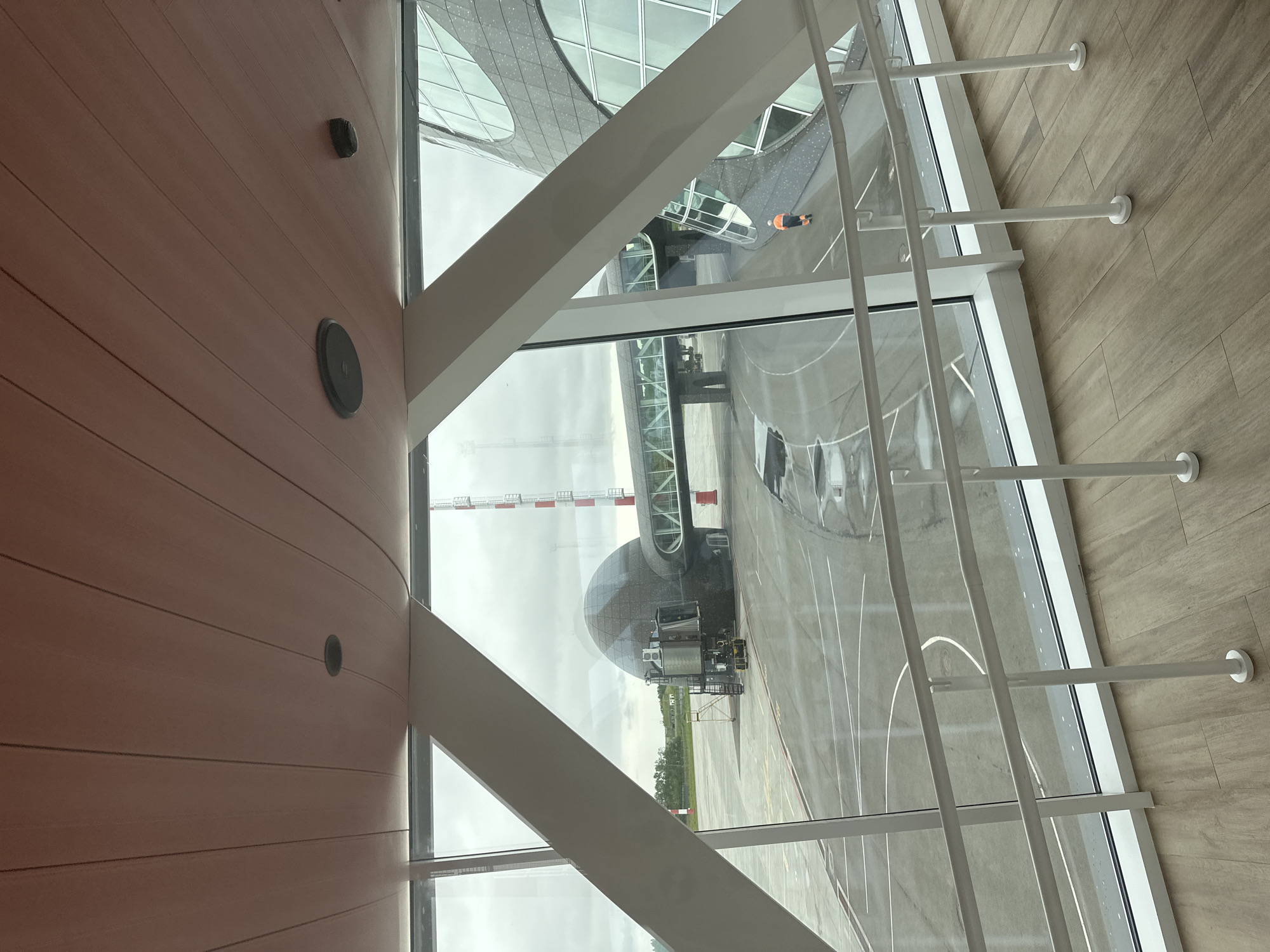 An airport jet bridge is attached to a rounded, dark-colored boarding structure, which is parked on an asphalt tarmac. The jet bridge extends from the structure towards a large, modern building with a glass facade. White handrails line a walkway in the foreground, and a person in an orange vest and dark pants walks away from the viewer on the tarmac. The sky is overcast, and the weather appears to be humid. The scene is likely at the Yuzhnyye Koryaki Airport in Yuzhnyye Koryaki, Russia.