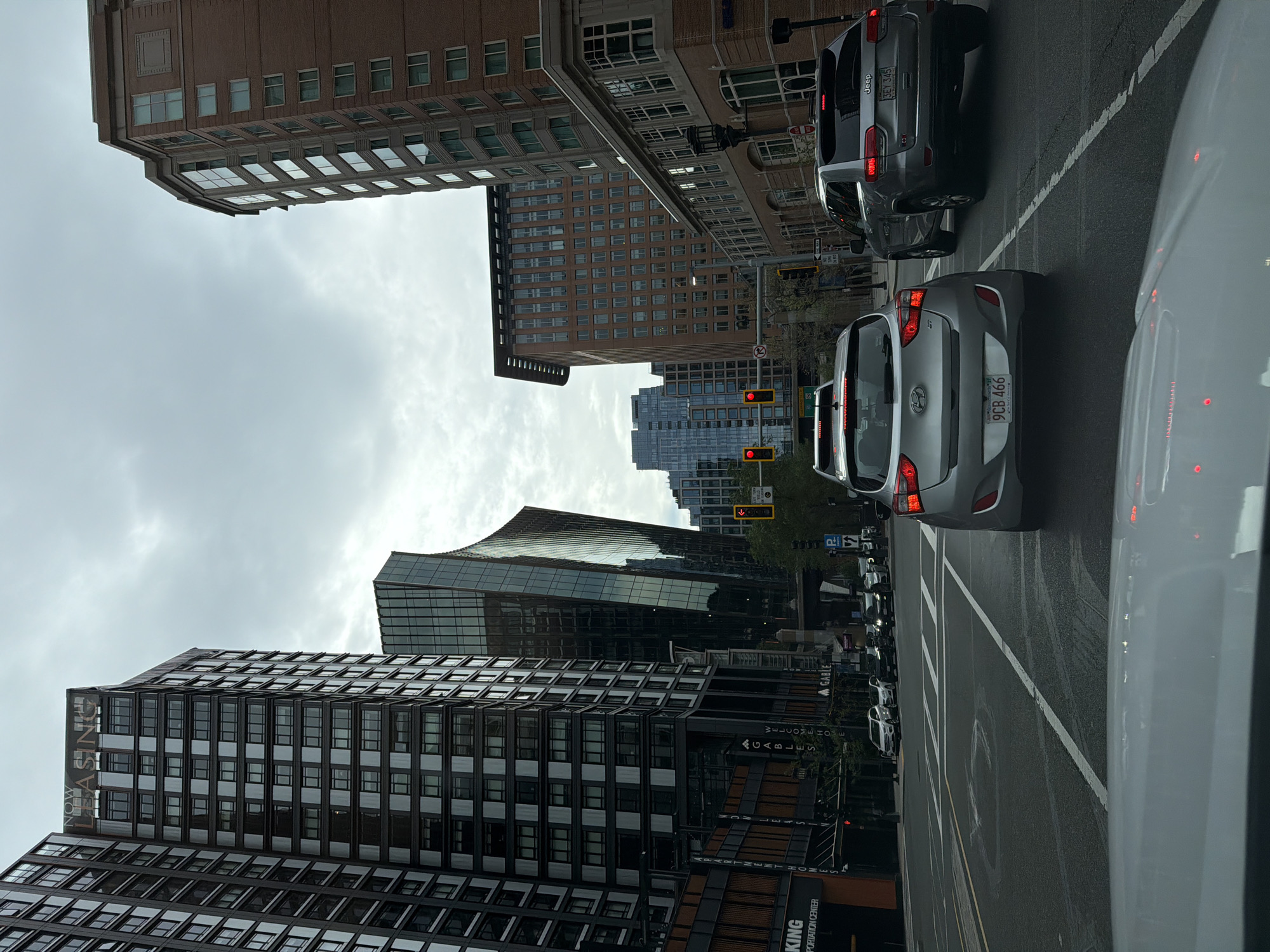 The image shows a street scene in Boston, Massachusetts, USA, on a cloudy day. In the foreground, a silver Hyundai hatchback is stopped at a traffic light. To its right, a silver Jeep Grand Cherokee is also stopped. The road is wet, indicating recent rain.
The background features several tall buildings, characteristic of a downtown urban environment. On the left, a dark-colored skyscraper with many windows dominates the view, with a sign that reads "NOW LEASING". To its right, a modern building with a distinctive curved, glass facade stands out. Further back, more contemporary buildings with reflective glass windows are visible.
Traffic lights are visible, with the red light illuminated, indicating that the vehicles are stopped. Street signs and lane markings are also present on the road. The overall impression is a typical city intersection with traffic and modern architecture.