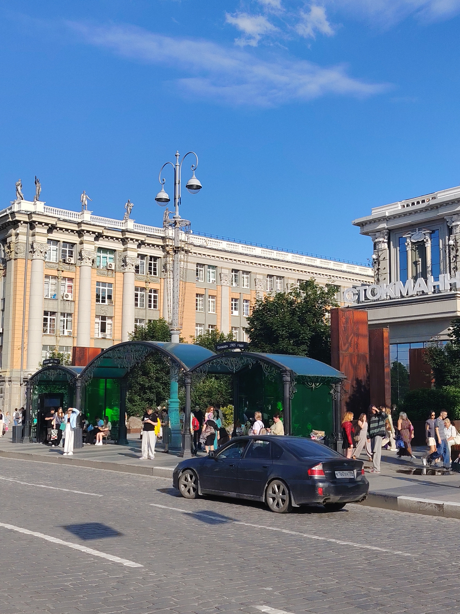 A sunny day is depicted in Yekaterinburg, Russia, with a clear blue sky and a few wispy clouds. The scene features a wide, cobblestone street in the foreground, with a dark sedan parked on the right side. White lines mark the lanes on the street.
Behind the street, a row of decorative green and black shelters with arched roofs are situated along a sidewalk. Several people are gathered around these shelters, some standing and some sitting. The shelters appear to be part of a public transport stop or a covered walkway.
Further back, a large, ornate building with a light orange and gray facade stands prominently. The building features classical architectural elements, including tall columns and statues adorning the roofline. To the right of this building is another modern structure with large windows and a prominent sign with white lettering.
The overall atmosphere is one of a bustling urban environment on a pleasant day. People are casually dressed, suggesting a leisurely activity or commute. The presence of architectural landmarks, public amenities, and pedestrians creates a vibrant city scene.
Visible text includes "ПЛОЩАДЬ 1905 ГОДА" on a sign under one of the shelters, and "СТОКМАНН" on the facade of the modern building. The license plate on the car reads "K160XH 198". The time appears to be late afternoon due to the long shadows cast by the buildings and streetlights.