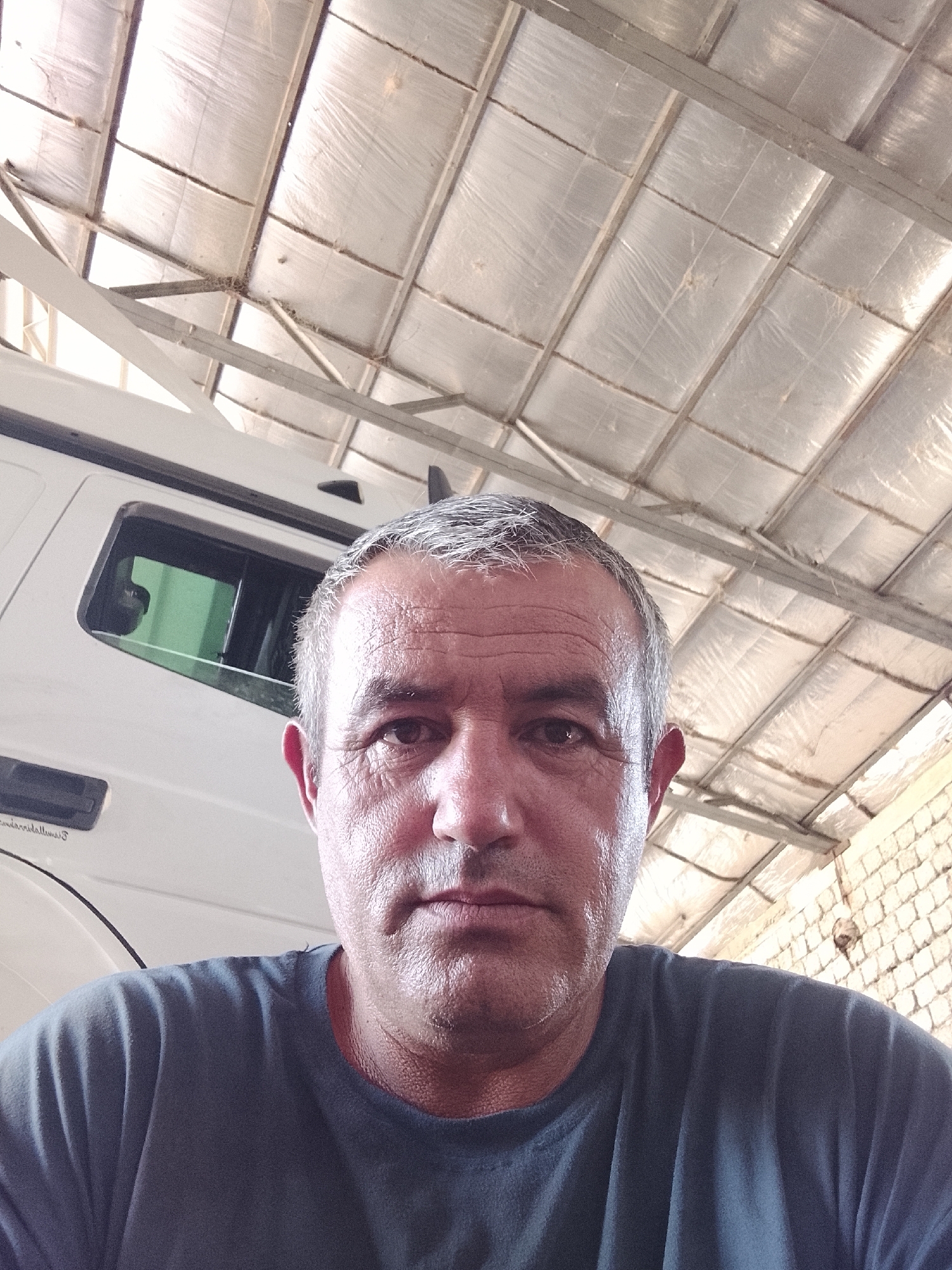 A medium shot photo shows a man looking directly at the camera. The man is wearing a dark blue t-shirt. He has short, graying hair and is looking straight ahead. Behind him, a white vehicle is visible, and above him is a metallic ceiling with rafters. The background is slightly out of focus. The image appears to have been taken indoors, possibly in a garage or workshop. There is no specific information to indicate the location in the city of Sabir Rakhimov, Uzbekistan.