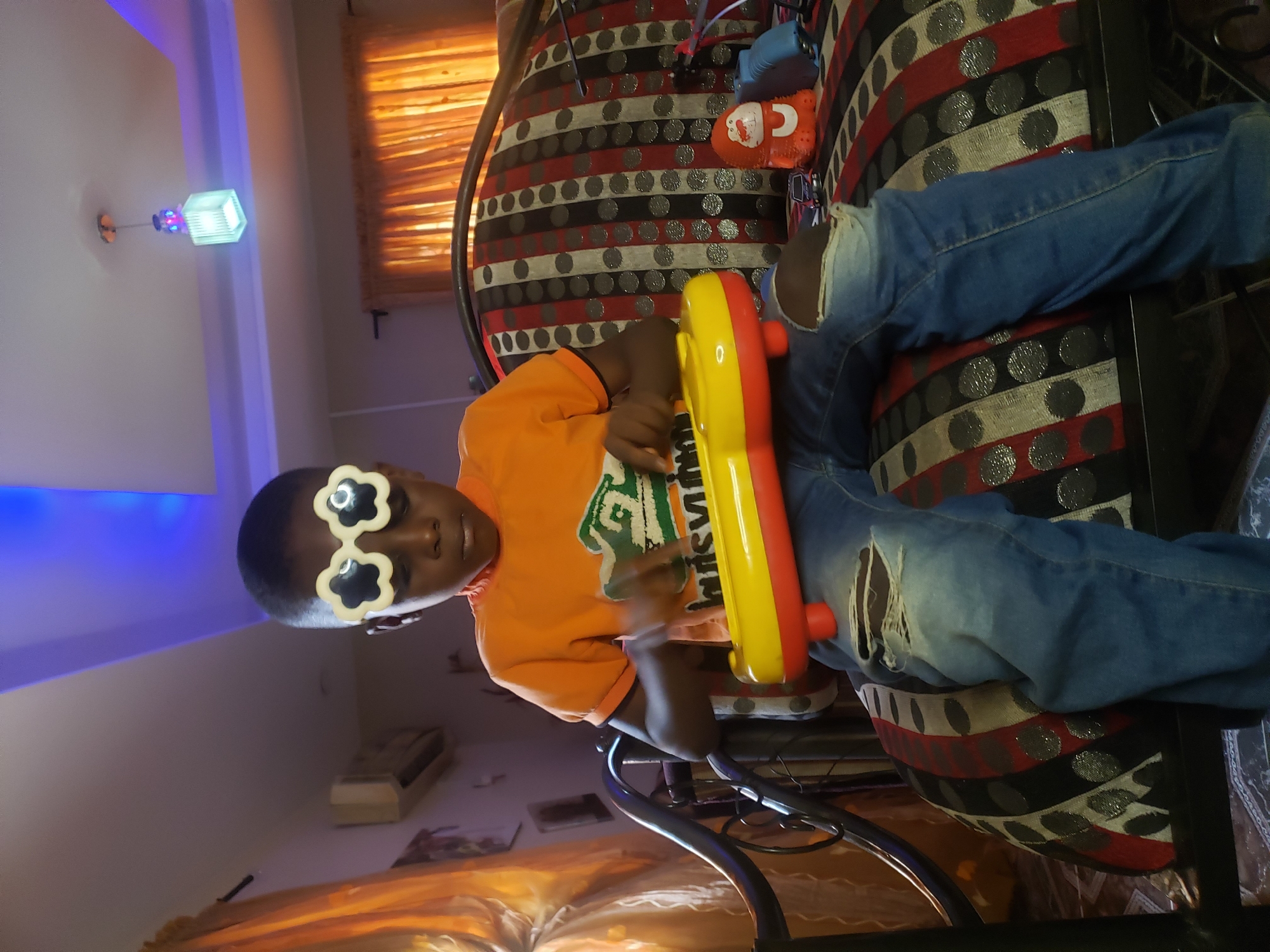 A young Black boy, wearing large, white, flower-shaped novelty glasses and an orange t-shirt with a green graphic, sits on a patterned sofa in Sabon Tasha, Nigeria. He is holding a yellow and red toy keyboard, with his hands positioned over the keys. The boy is wearing distressed blue jeans with rips on the knees. The sofa has a black, white, red, and grey striped pattern with circular accents. Next to him on the sofa, there is a small orange monkey-like toy and a blue toy. In the background, orange curtains are visible, along with an air conditioning unit mounted on the wall. A ceiling light fixture hangs from the ceiling, which is illuminated with blue LED lights. The room appears to be a living room.