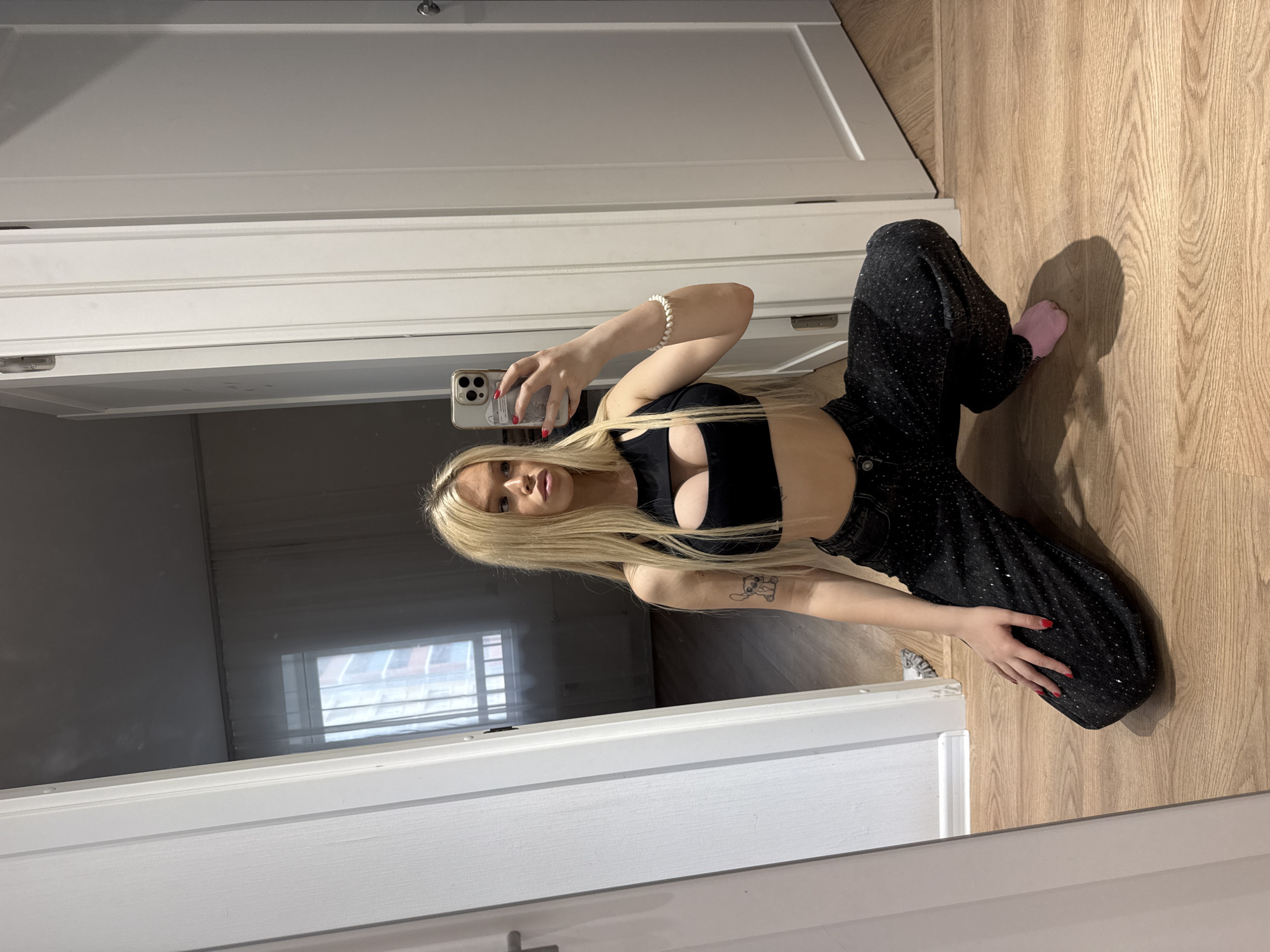 A young woman with long blonde hair and heavy makeup is seen from the waist up in a mirror reflection, kneeling on a wooden floor. She is wearing a black crop top with a cutout in the chest area and black, wide-leg jeans adorned with rhinestones. On her left arm, she has a tattoo of the character Stitch. Her right hand is holding a smartphone, presumably to take the picture. She is also wearing a white beaded bracelet on her right wrist and a pink sock on her left foot. The background shows a doorway with white doors and a window with sheer curtains, suggesting an indoor setting, possibly an apartment. The floor is a light-colored wood. The image is taken indoors, reflecting off a mirror. The context of Petro-Slavyanka, Russia, is not discernible from the visual information provided in the image itself.